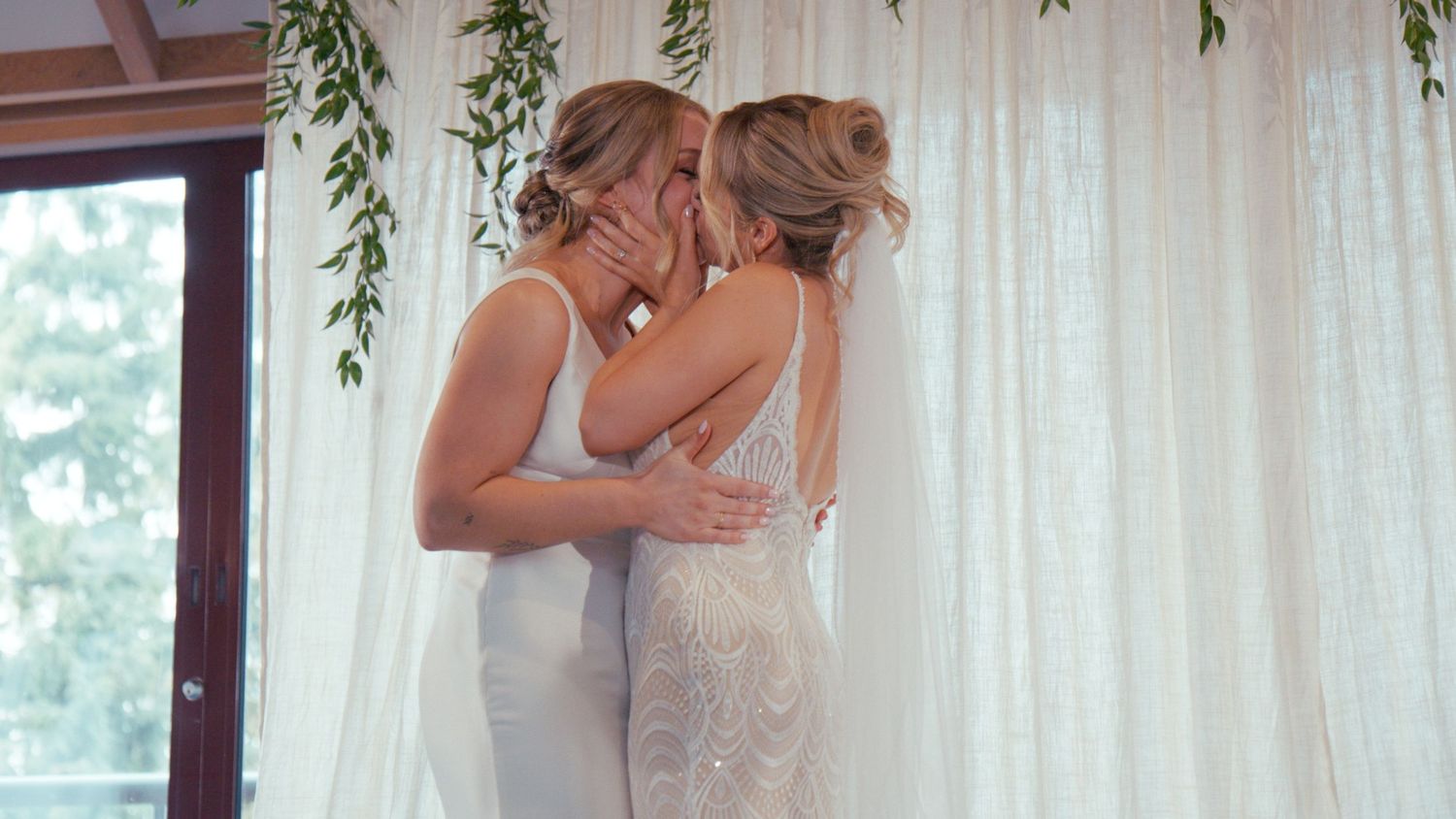 Two brides share a romantic kiss in front of white curtain backdrop with hanging greenery.