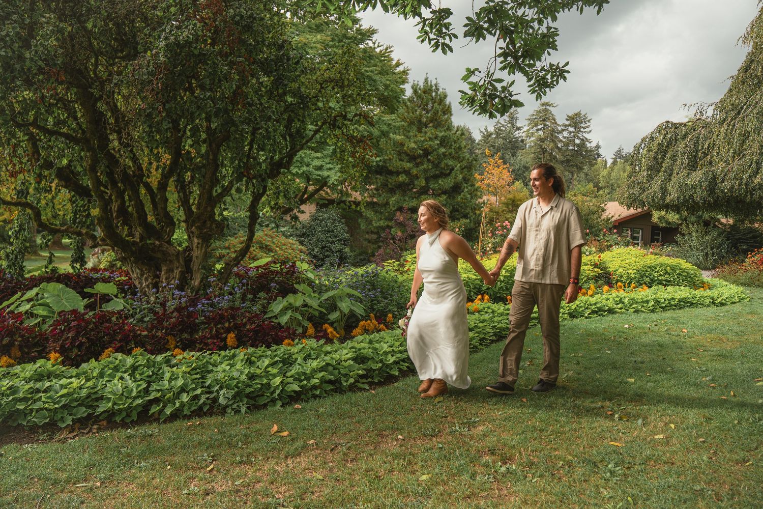 Couple holding hands walking through lush garden with colorful flowers and trees on a cloudy day.