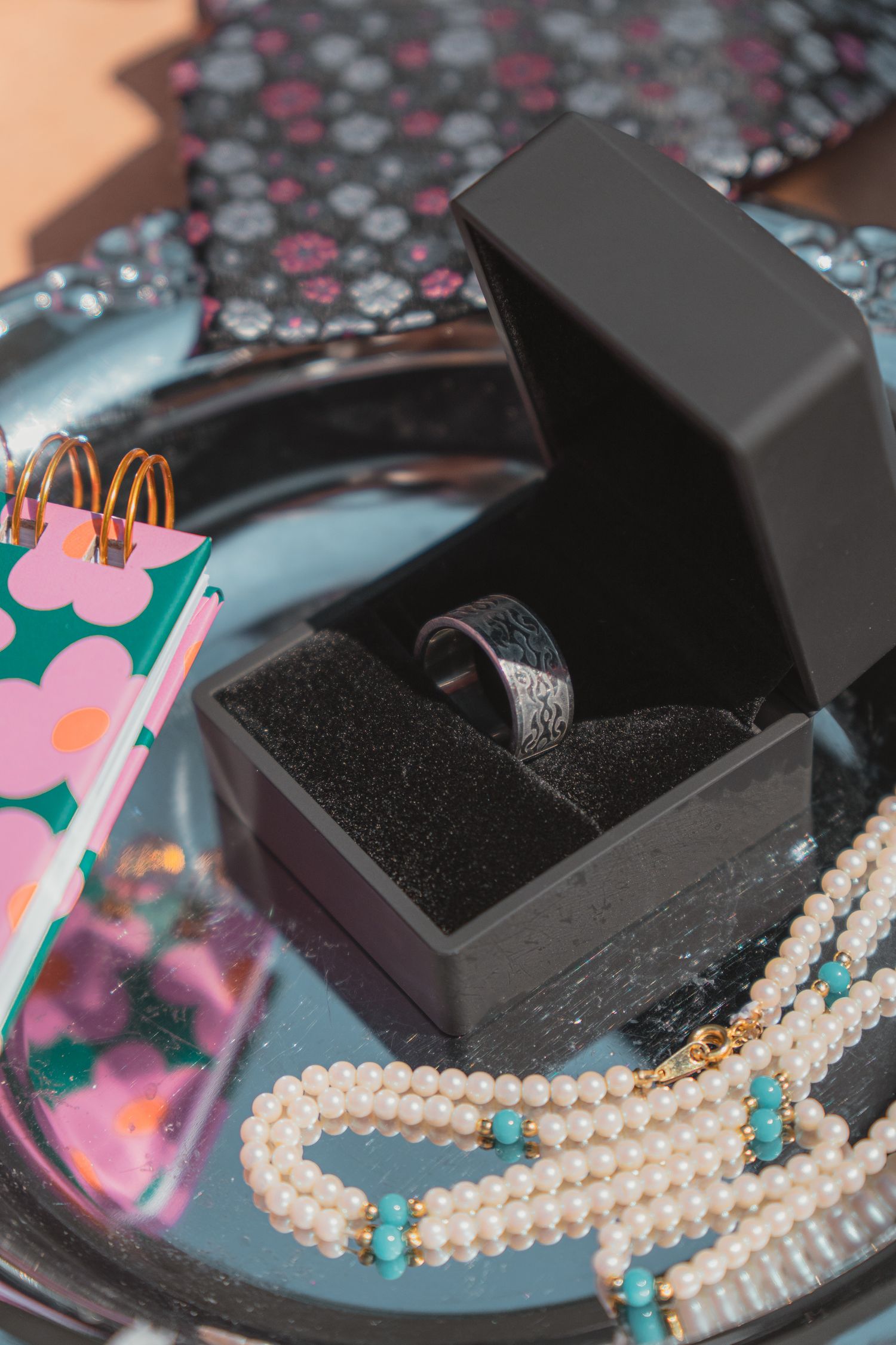 Black jewelry box with silver ring, pearl necklace with turquoise beads, and floral gift bag on a dark tray.