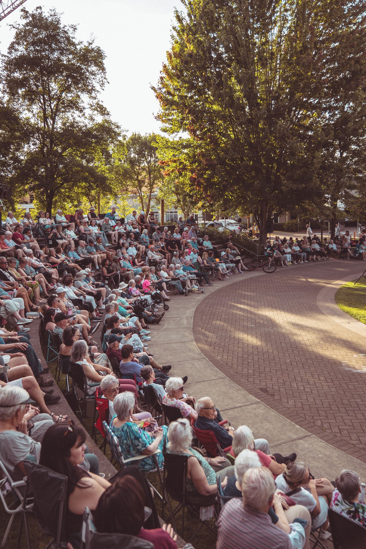 Large outdoor audience seated in curved rows at a sunny park amphitheater surrounded by tall green trees.