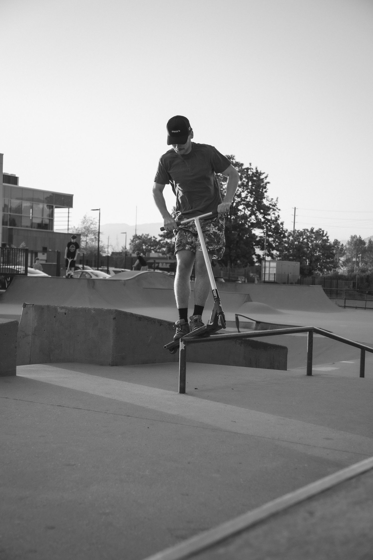 Black and white photo of a man balancing on a scooter over a rail at a skate park.