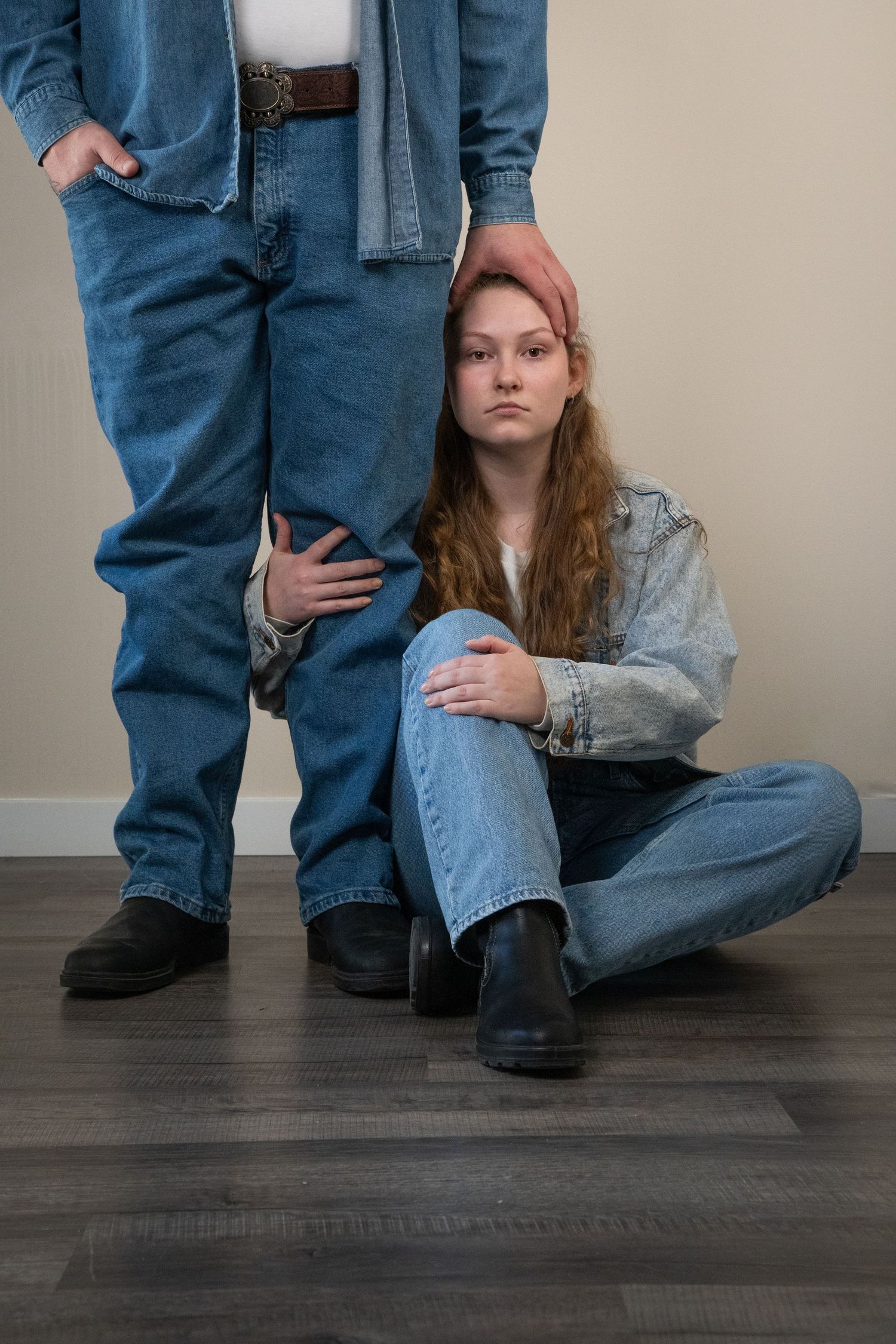Young woman in denim jacket sitting on floor leaning against standing mans legs, indoor portrait session.
