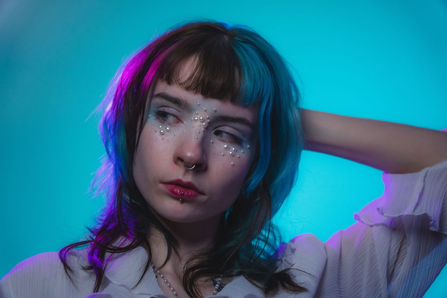 Woman with teal hair and pearl face gems posing under neon blue and pink studio lighting.