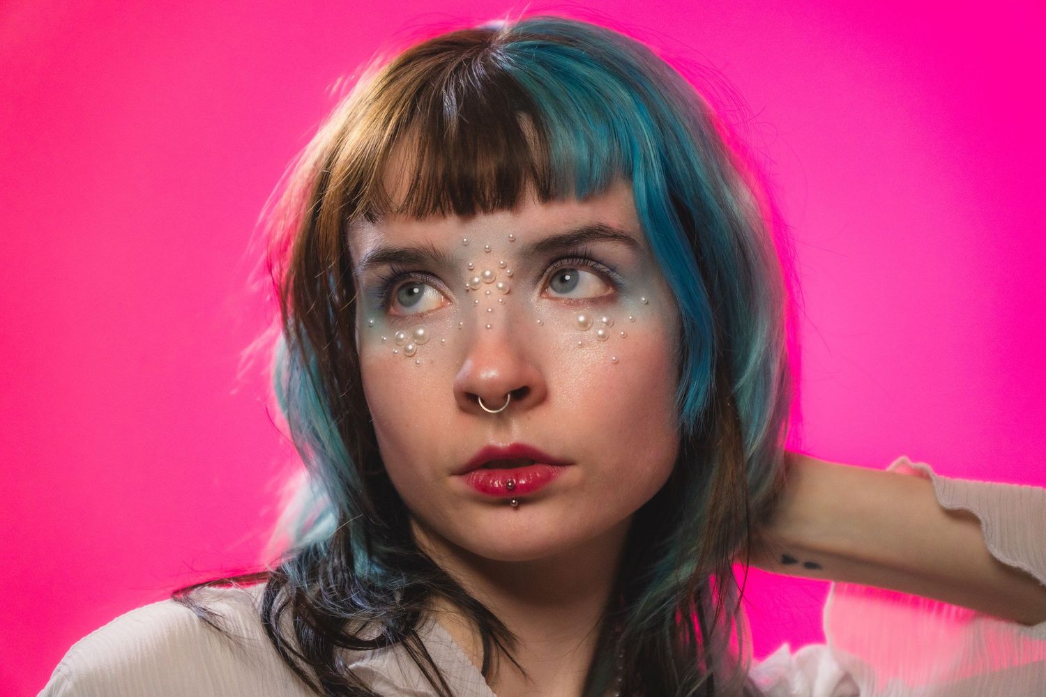 Woman with teal hair, nose ring, and glitter gems looking sideways on pink background.