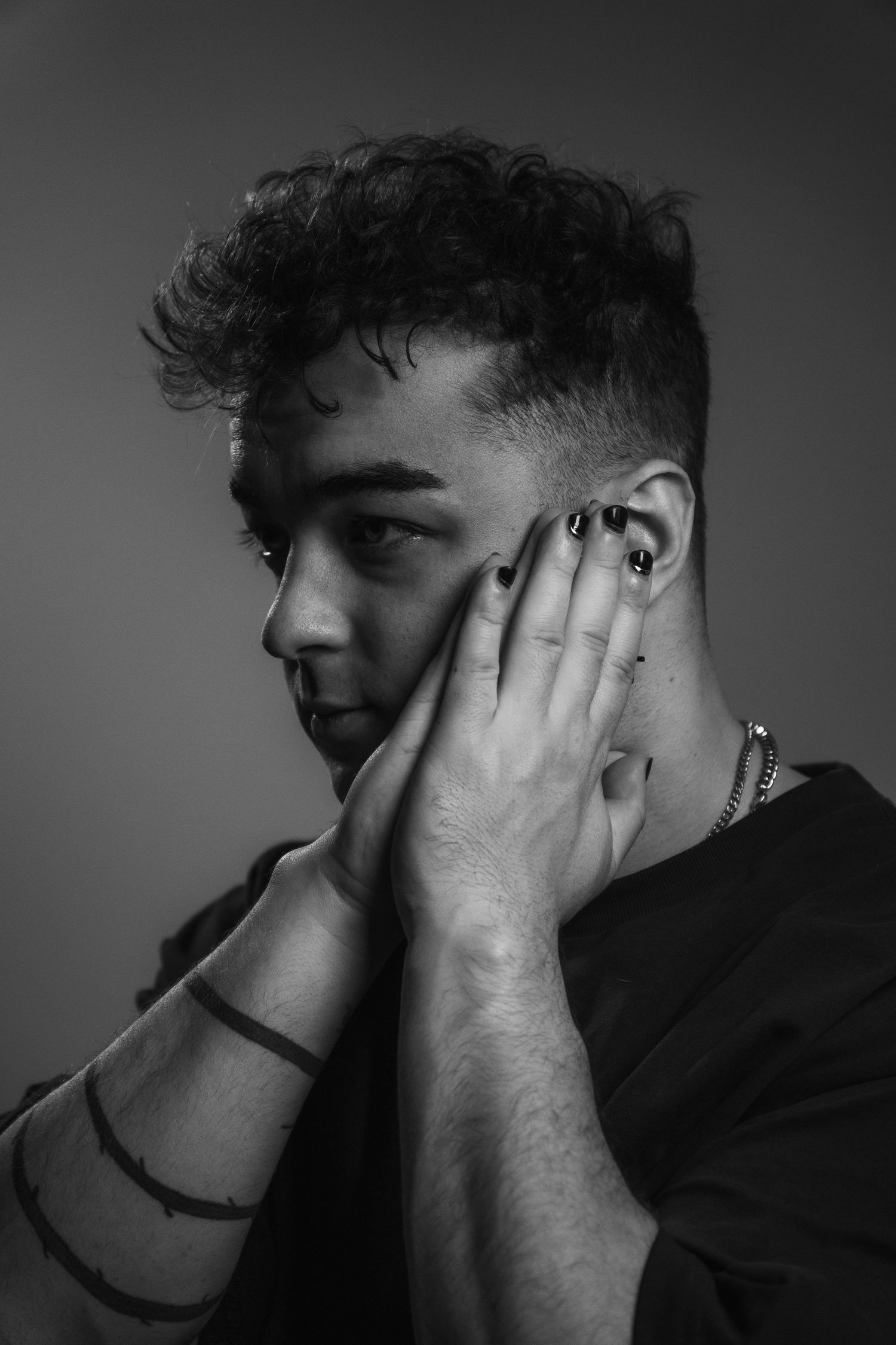Black and white close-up of young man with curly hair holding face with both hands, wearing bracelets.