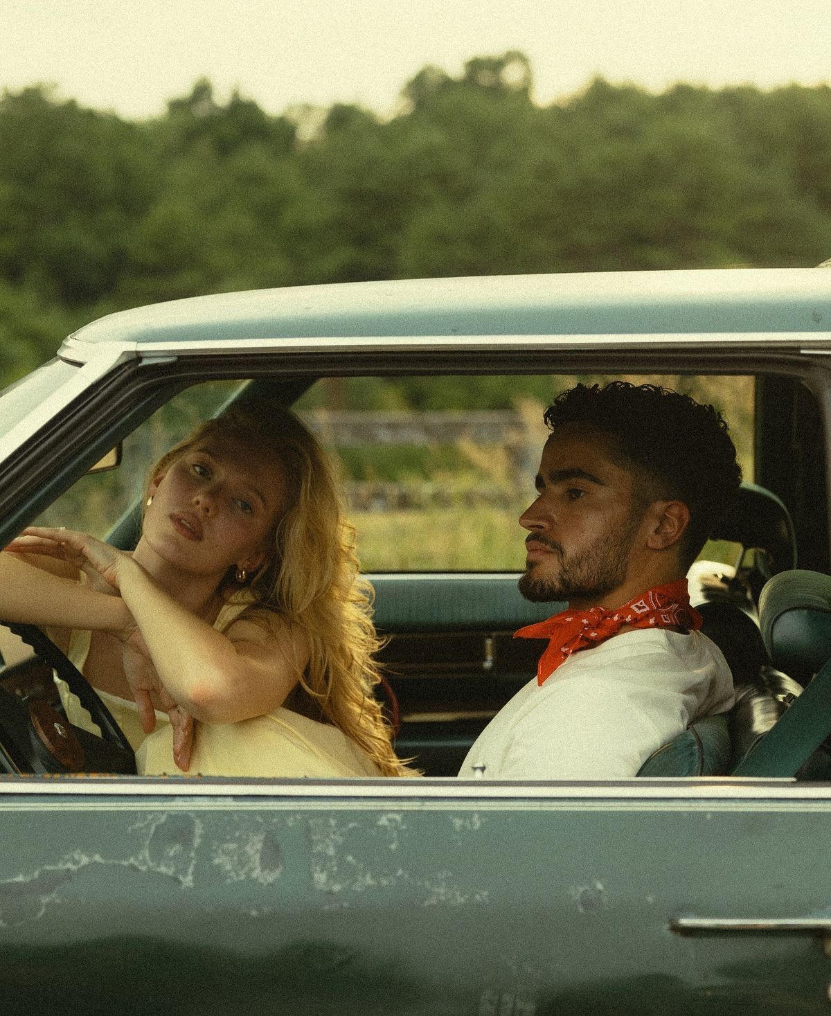 A young couple sits in a vintage teal car, woman with blonde hair in yellow dress, man wearing red bandana.