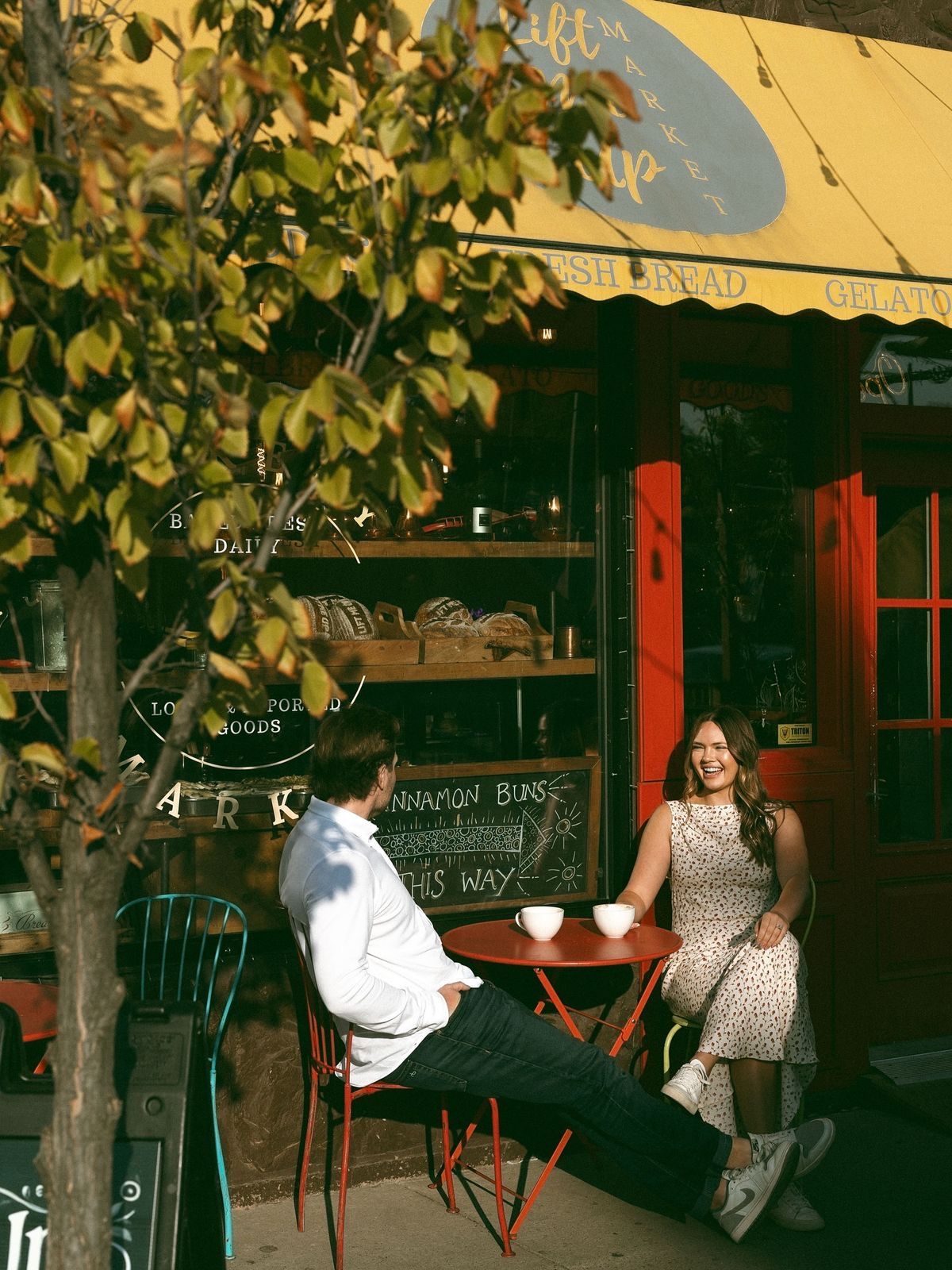 Couple enjoying coffee at outdoor cafe table beside a yellow-awning bakery storefront with lemon tree.