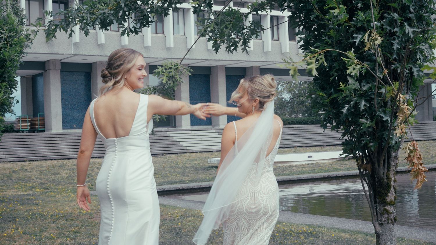 Two brides in white wedding dresses dancing together outdoors near a fountain.