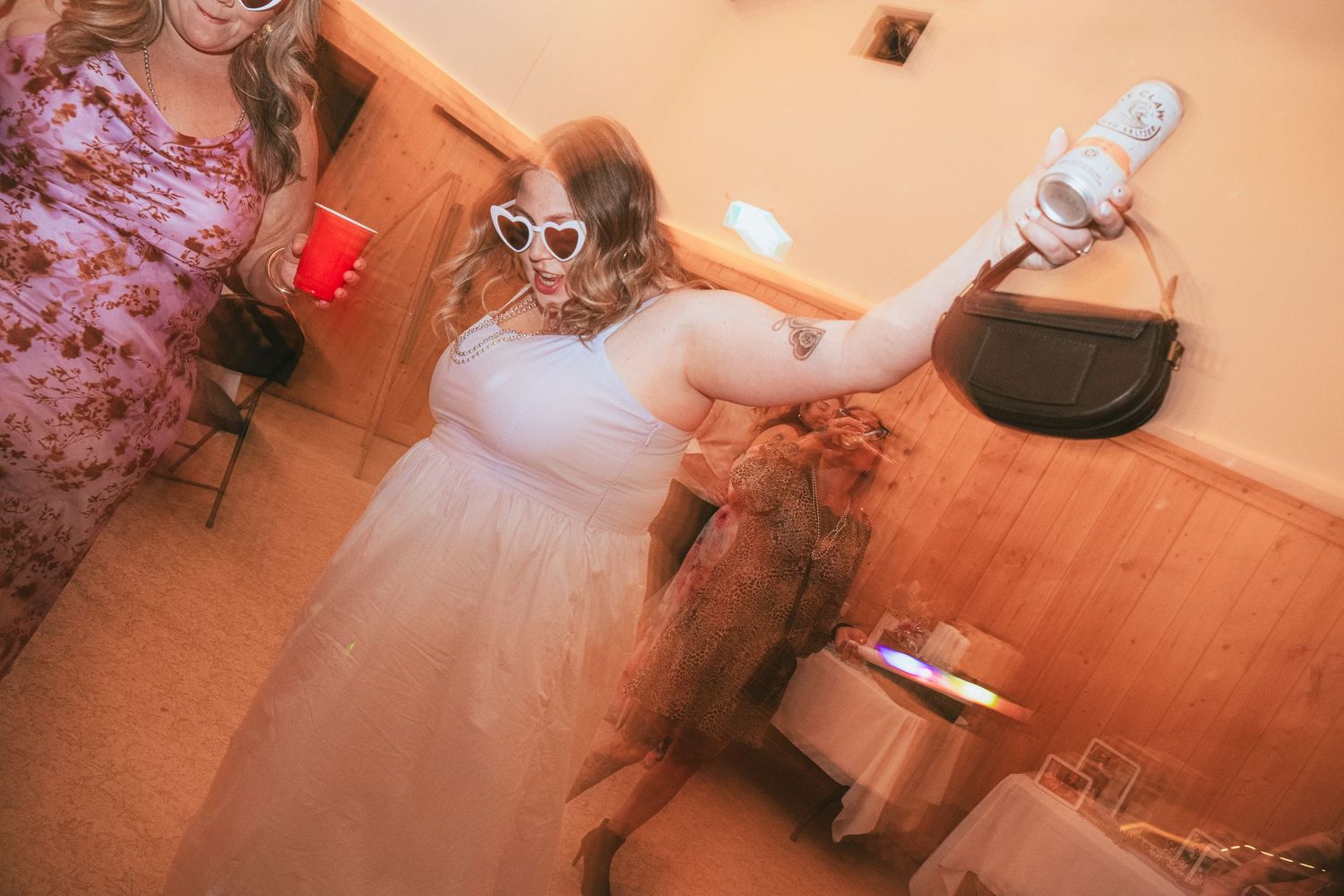 A bride in a white tulle skirt and heart sunglasses takes a selfie while dancing with a dog at a party.
