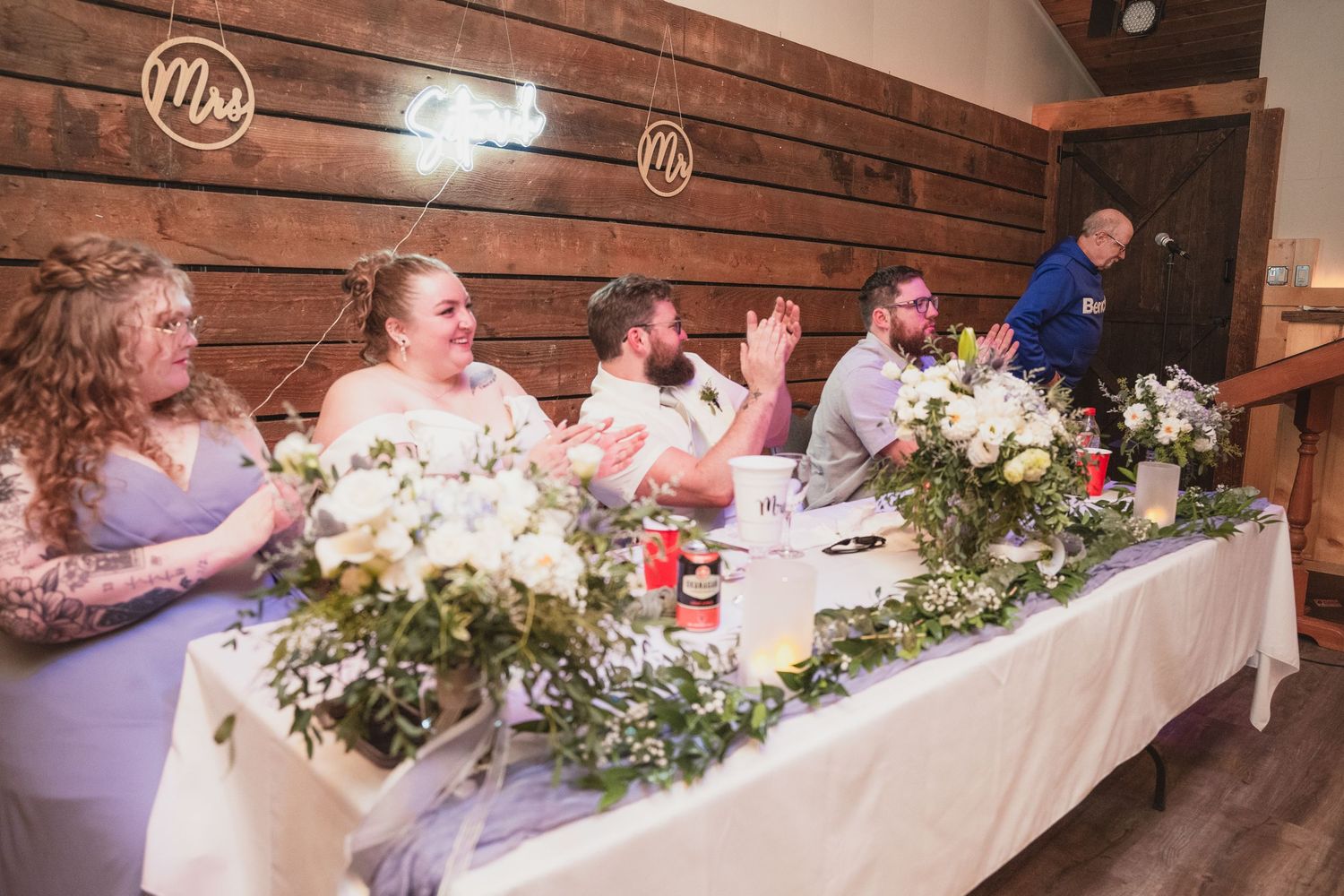 Wedding party seated at decorated head table with floral arrangements during barn reception speeches.