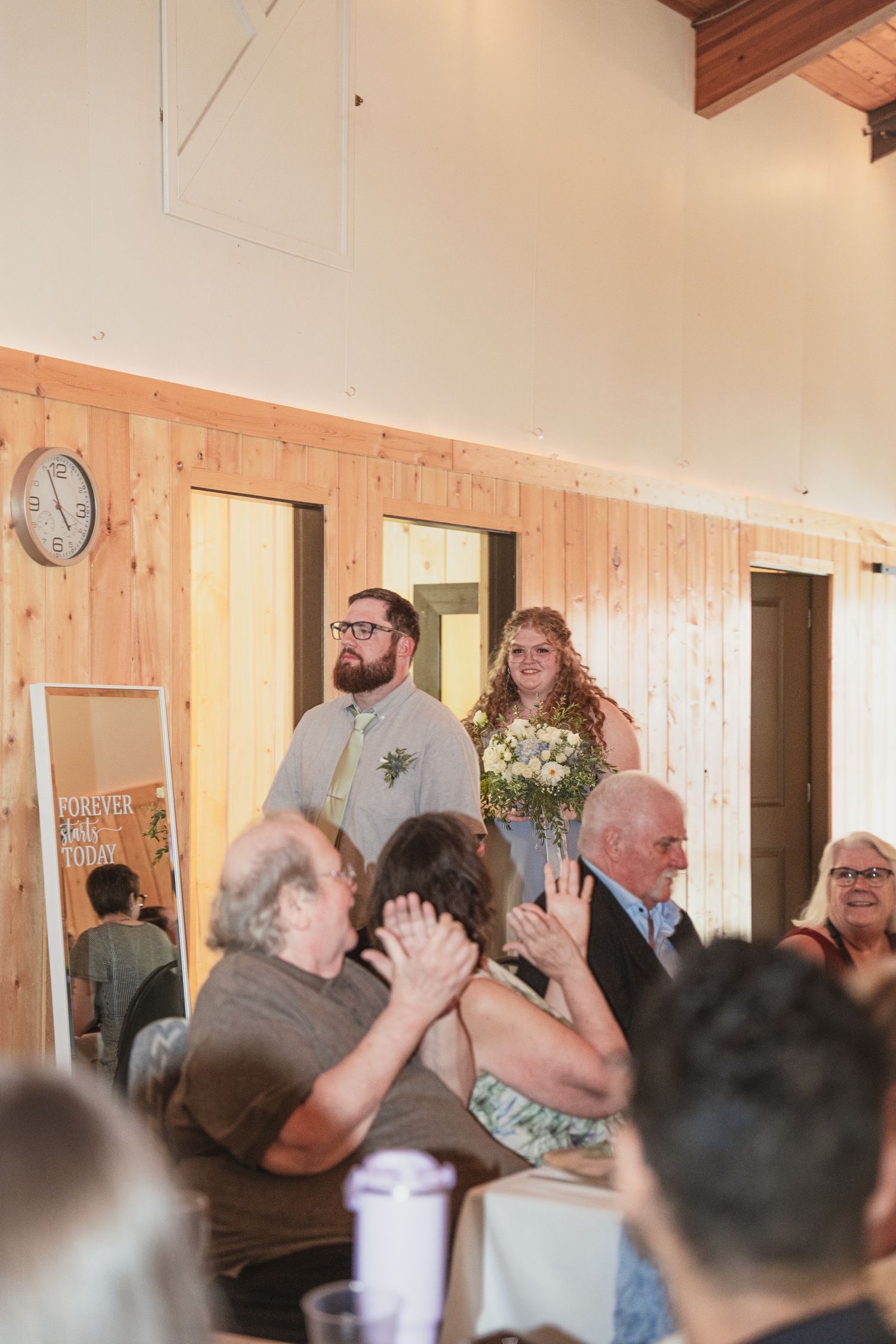 Bride and groom enter wedding reception hall greeted by applauding guests in a rustic wood-paneled venue.