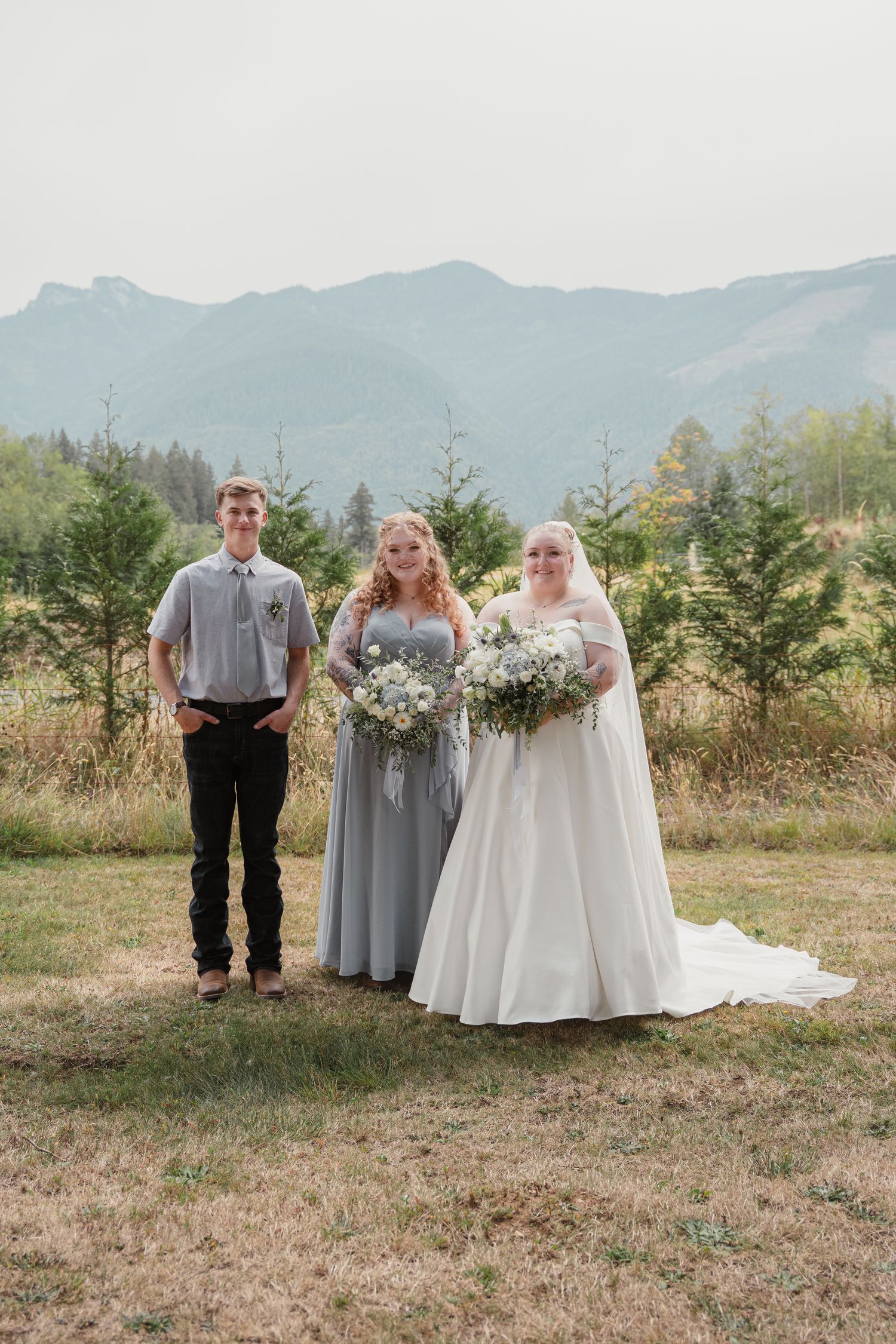 Bride with bridesmaid in gray dress and young man posing outdoors with mountain backdrop.