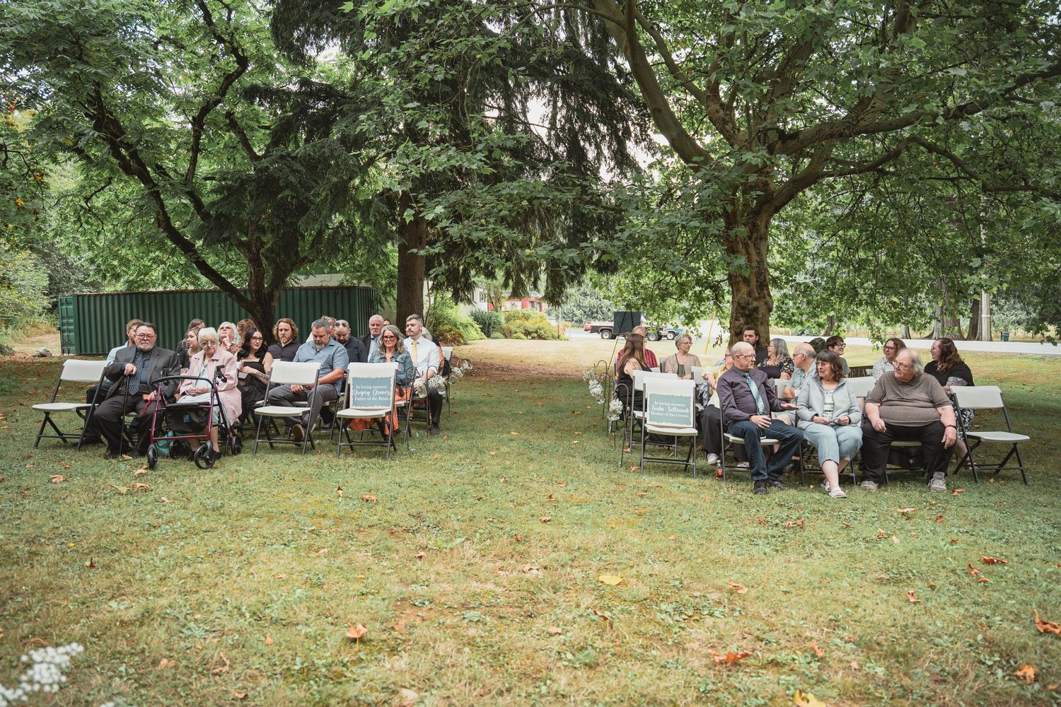 Outdoor wedding ceremony with guests seated on chairs under large trees in a lush green park setting.