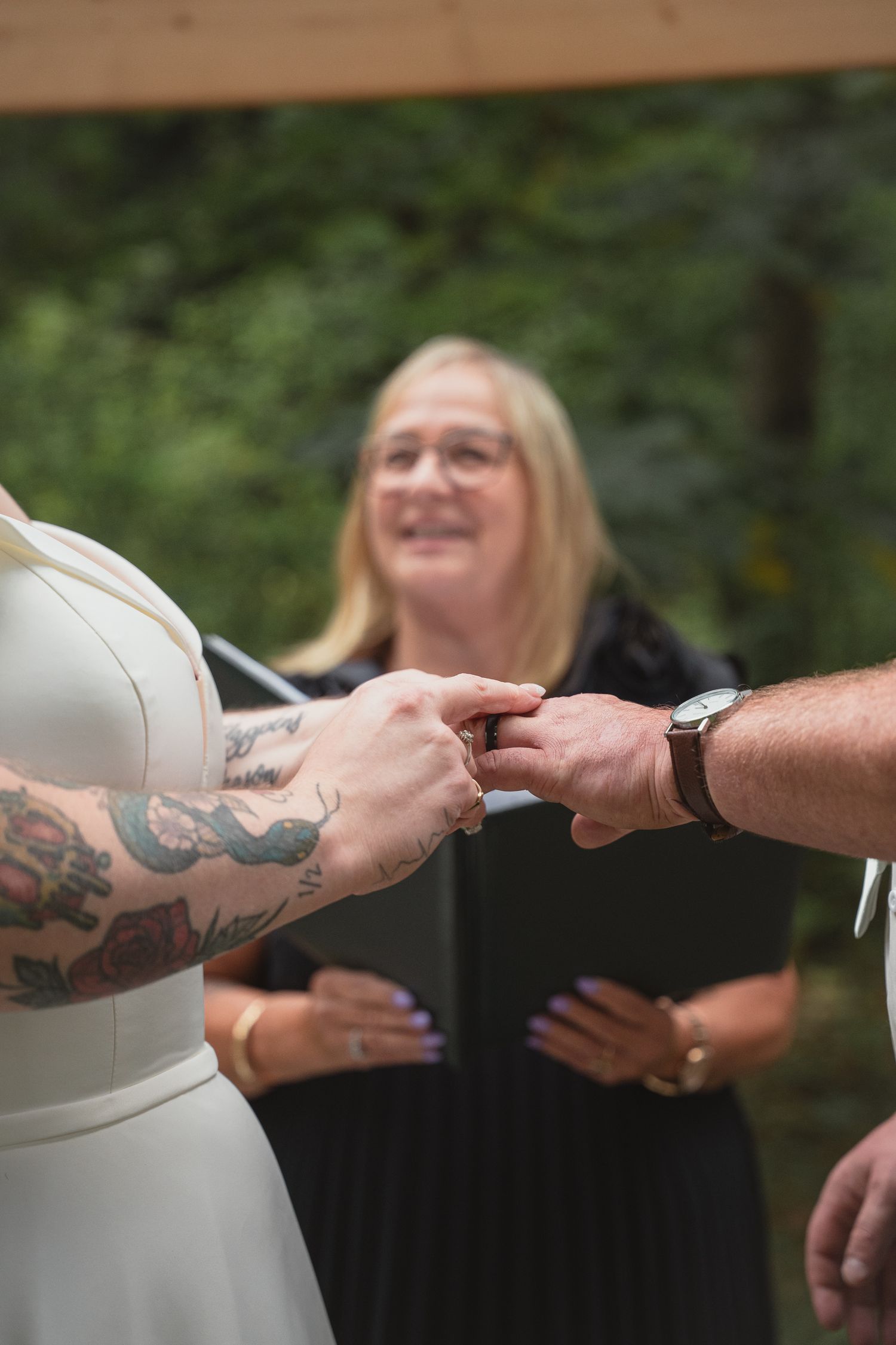 Couple exchanging rings during outdoor wedding ceremony with smiling officiant in black robe standing behind them.