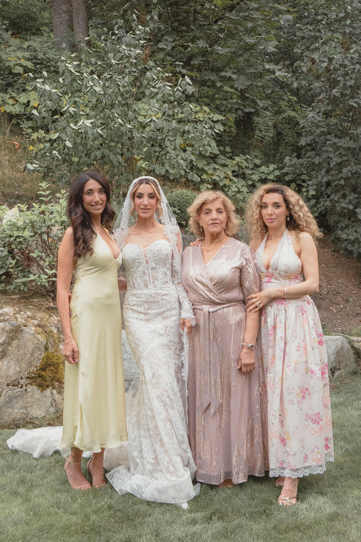 Bride in lace gown with veil poses alongside three women in elegant dresses outdoors at a garden wedding.