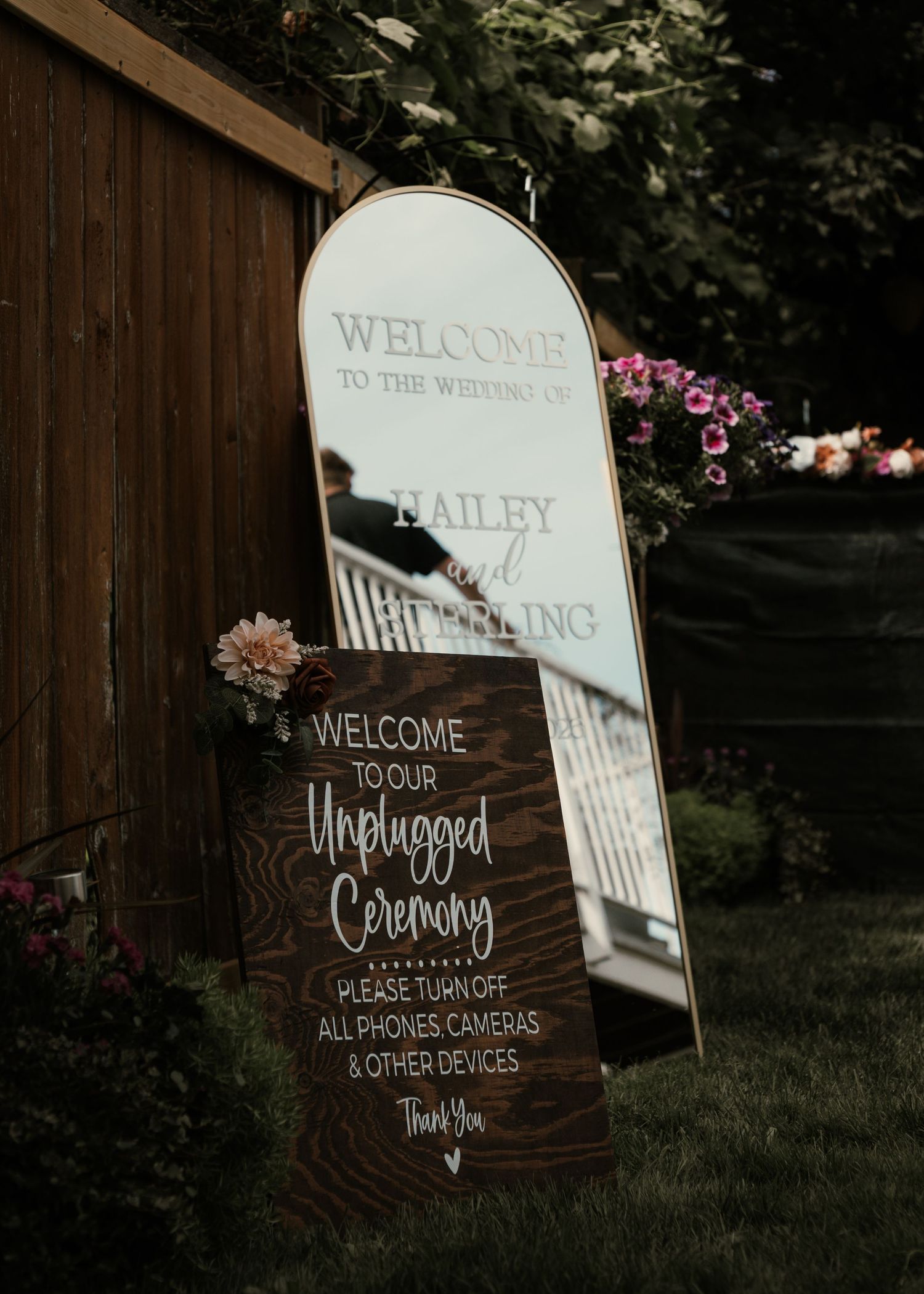 Rustic wooden unplugged ceremony wedding sign beside an arched mirror welcome sign in an outdoor garden setting.