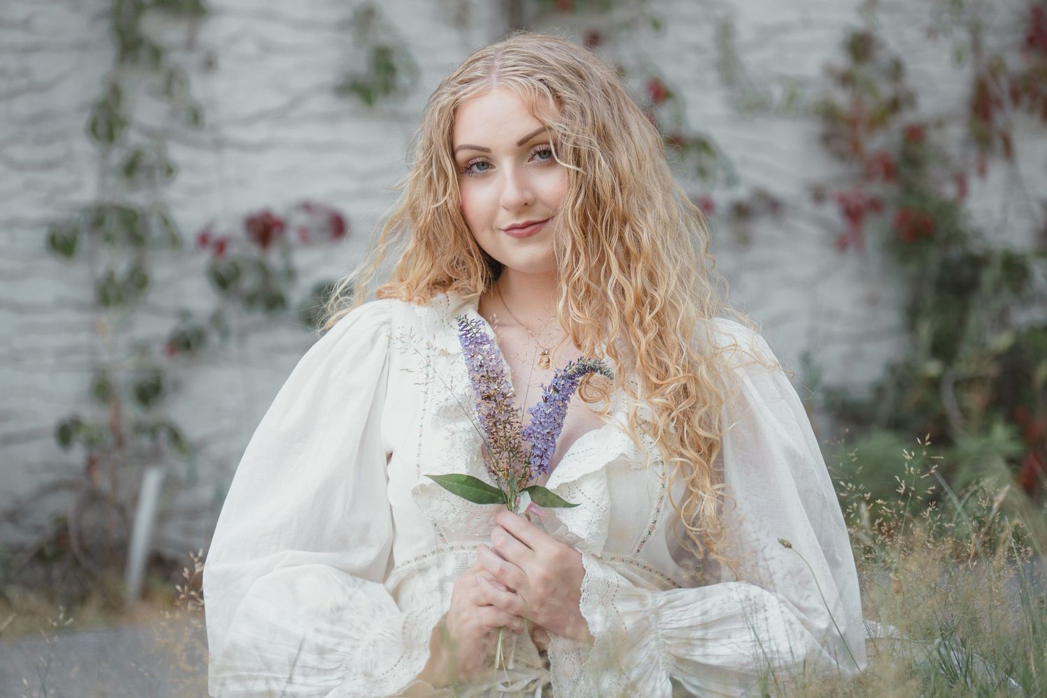 Young woman with curly blonde hair in white Victorian dress holding purple wildflowers outdoors.