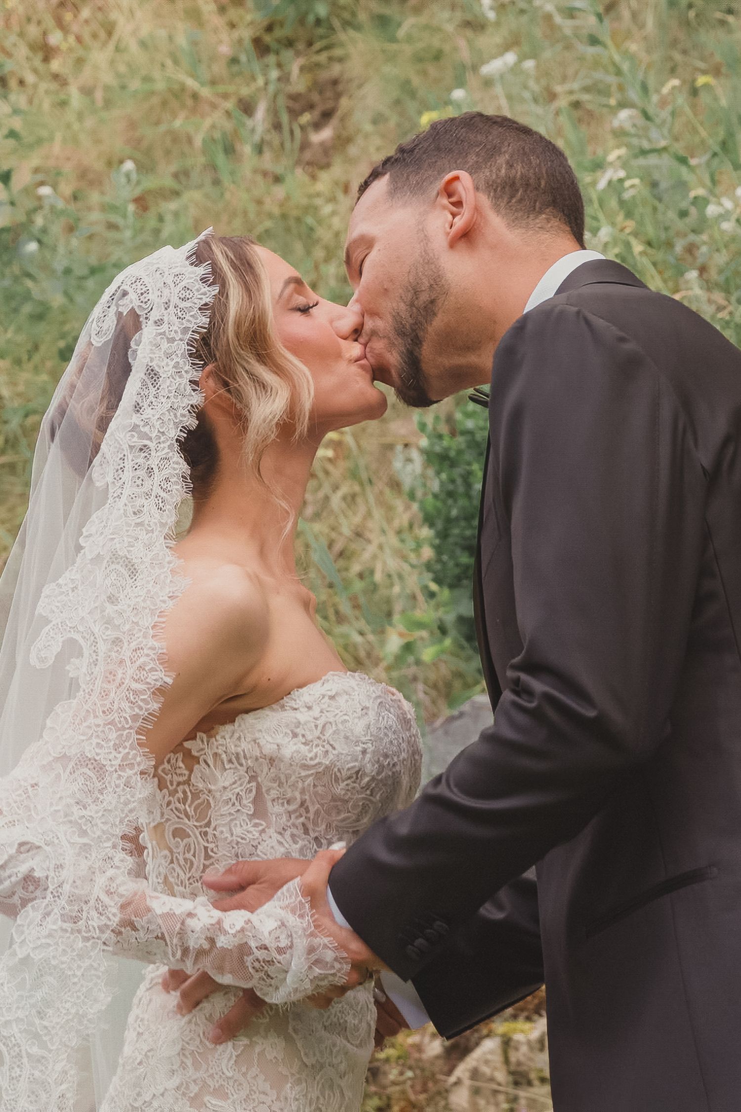 Bride and groom sharing a romantic kiss outdoors, bride wearing lace wedding dress and veil, groom in dark suit.