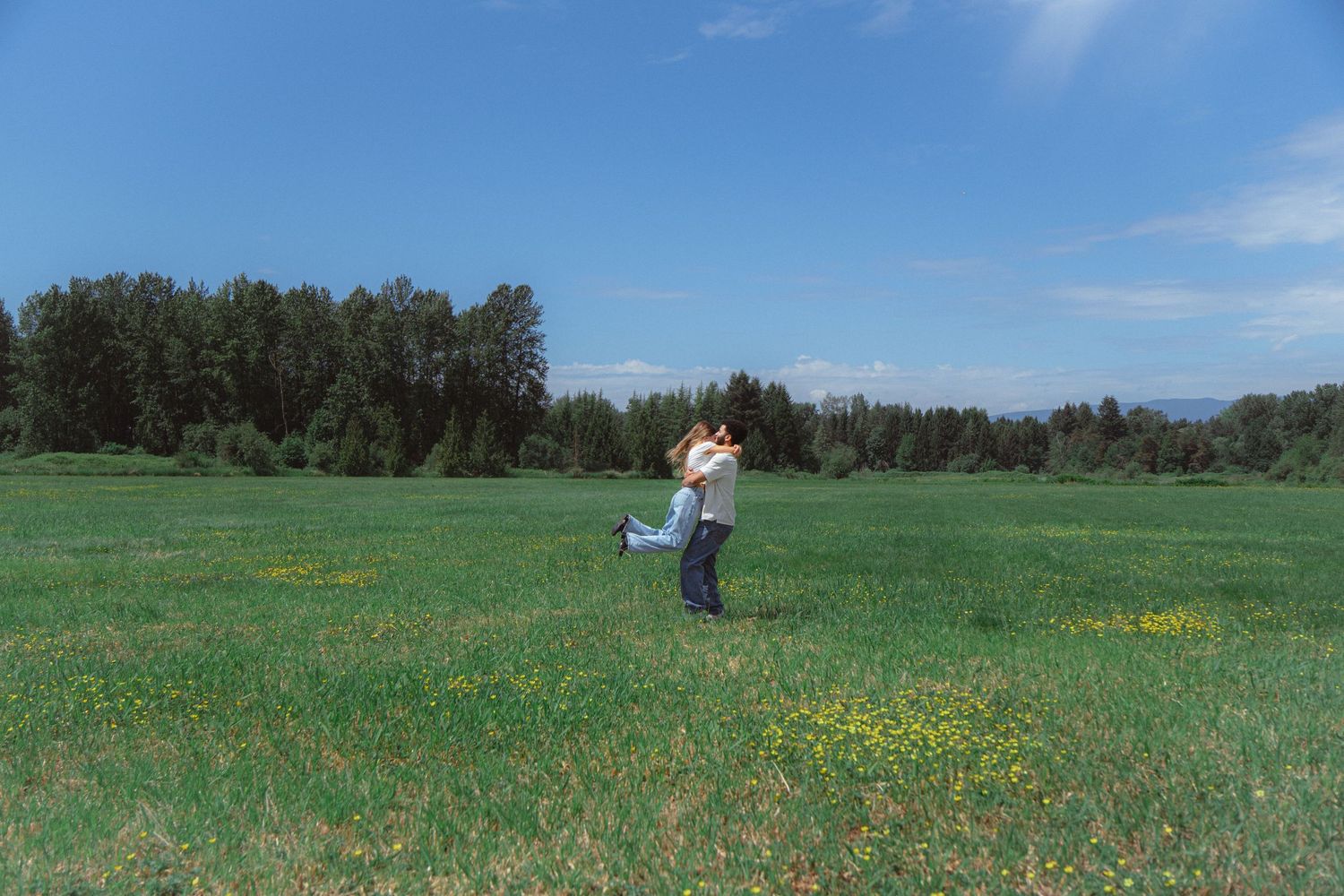 A couple embraces and spins in a vast green meadow under a bright blue sky.