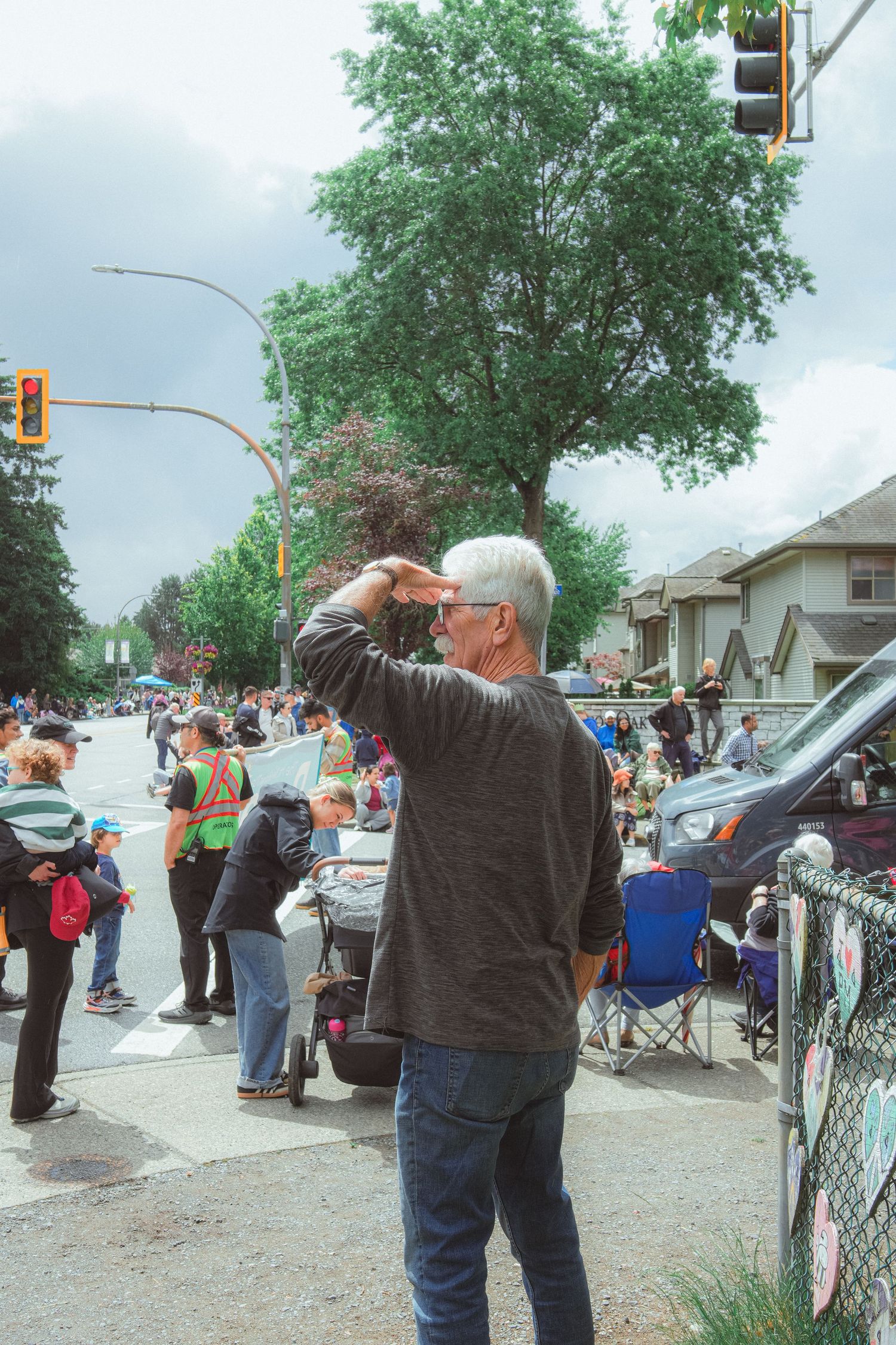 Silver-haired man shields his eyes watching a busy street parade with spectators lining the sidewalk.