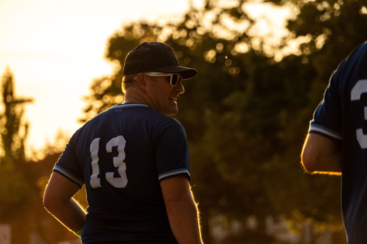 Softball player wearing number 13 Dodgers jersey stands in warm sunset light during an evening game.