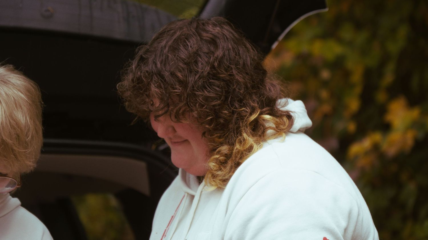 Person with curly brown hair wearing a white hoodie stands outdoors near a vehicle in autumn.
