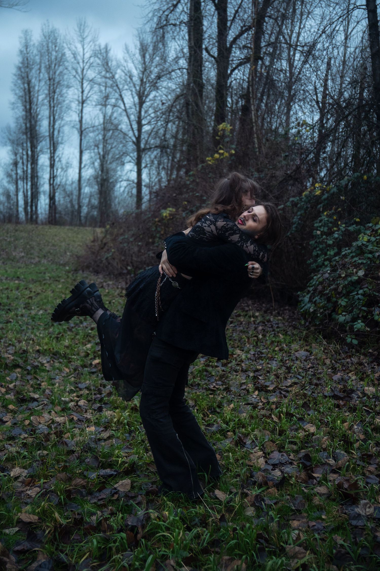 Gothic man lifts woman in black outfit on a leaf-covered ground near bare trees.