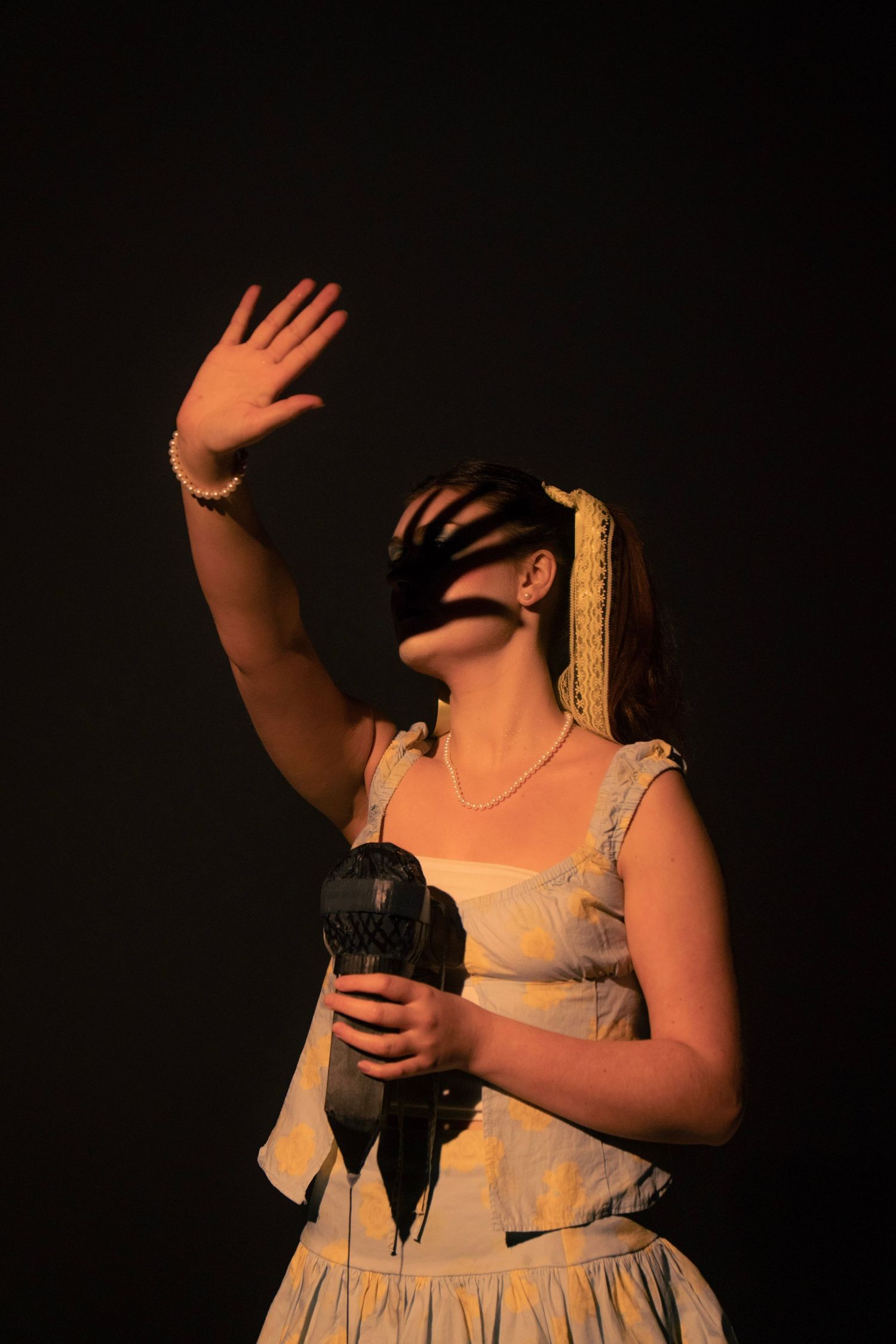 Woman in vintage dress holding microphone with raised hand, dramatic shadows across face on dark background.