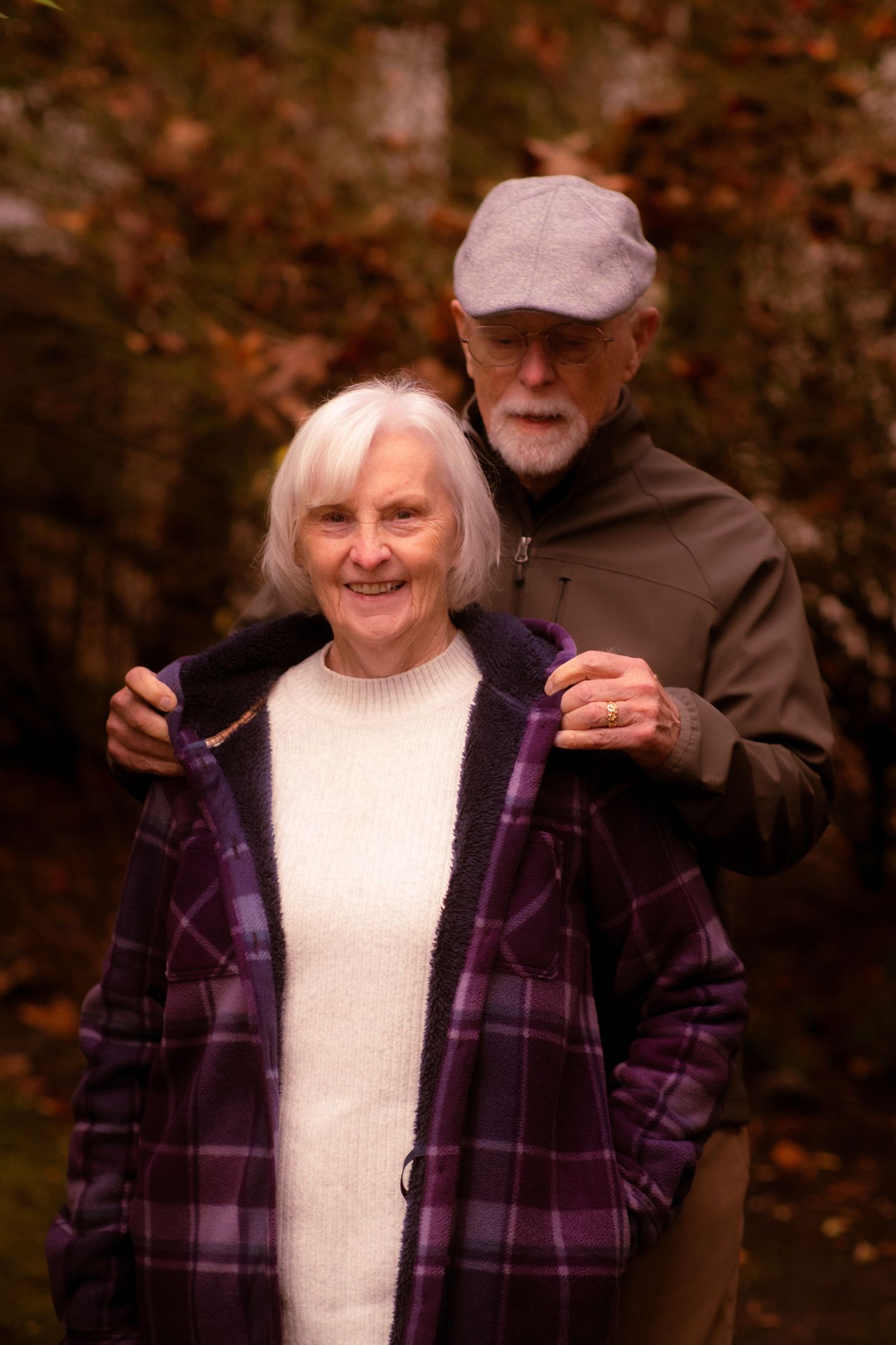 Senior man lovingly helping woman put on purple plaid coat outdoors during autumn season.