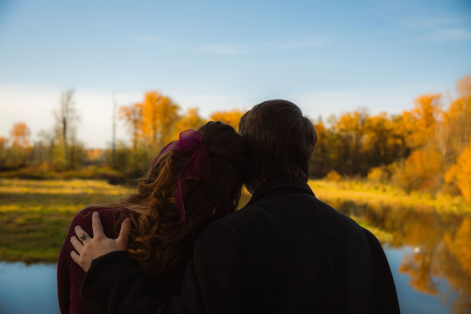 Couple viewed from behind sharing a tender moment by a calm lake surrounded by vibrant autumn foliage.