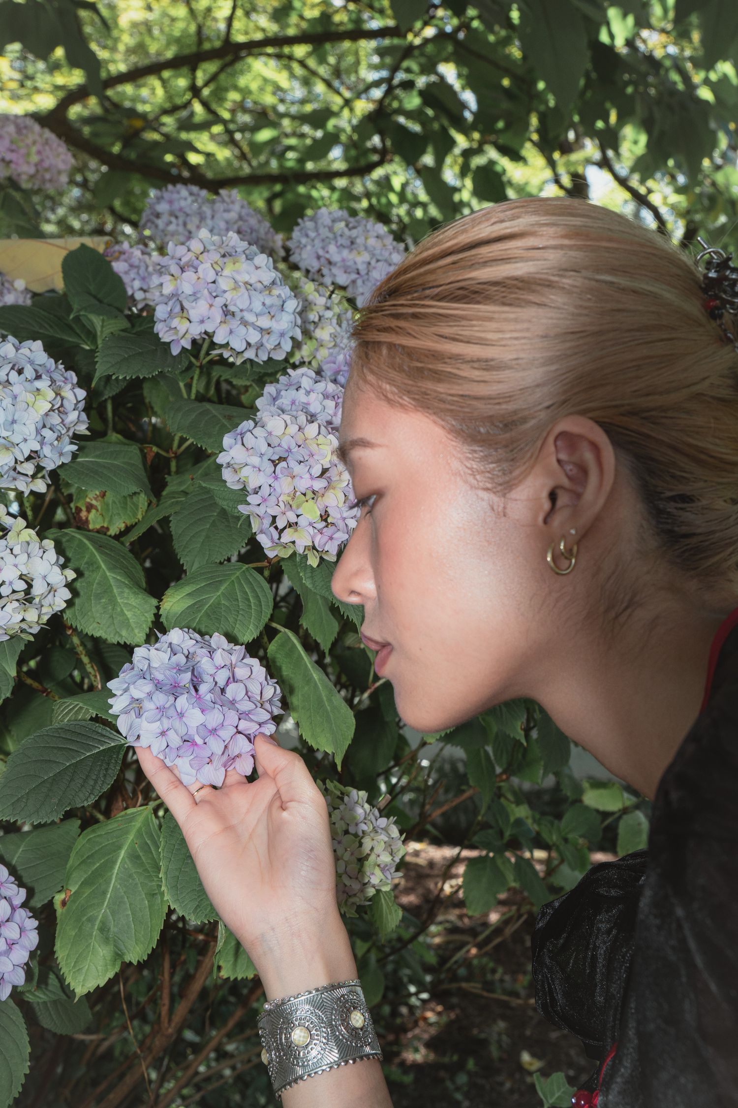 Young blonde woman smelling purple hydrangea flowers in a lush green garden, wearing a silver cuff bracelet.