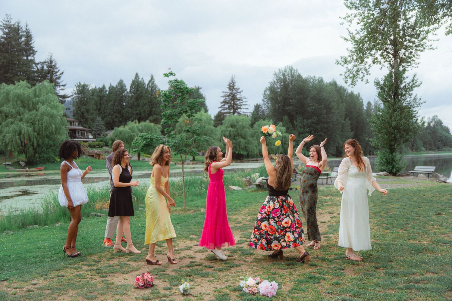 Bride tossing bridal bouquet to eager bridesmaids in colorful dresses at outdoor lakeside wedding.