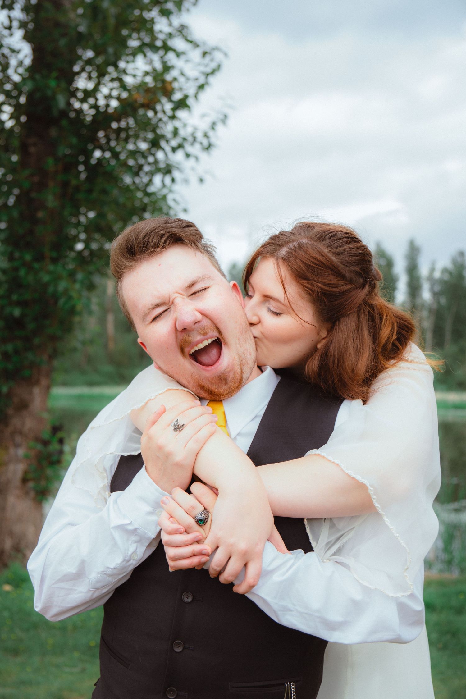 Bride playfully kisses laughing groom from behind in a fun outdoor wedding portrait near trees.