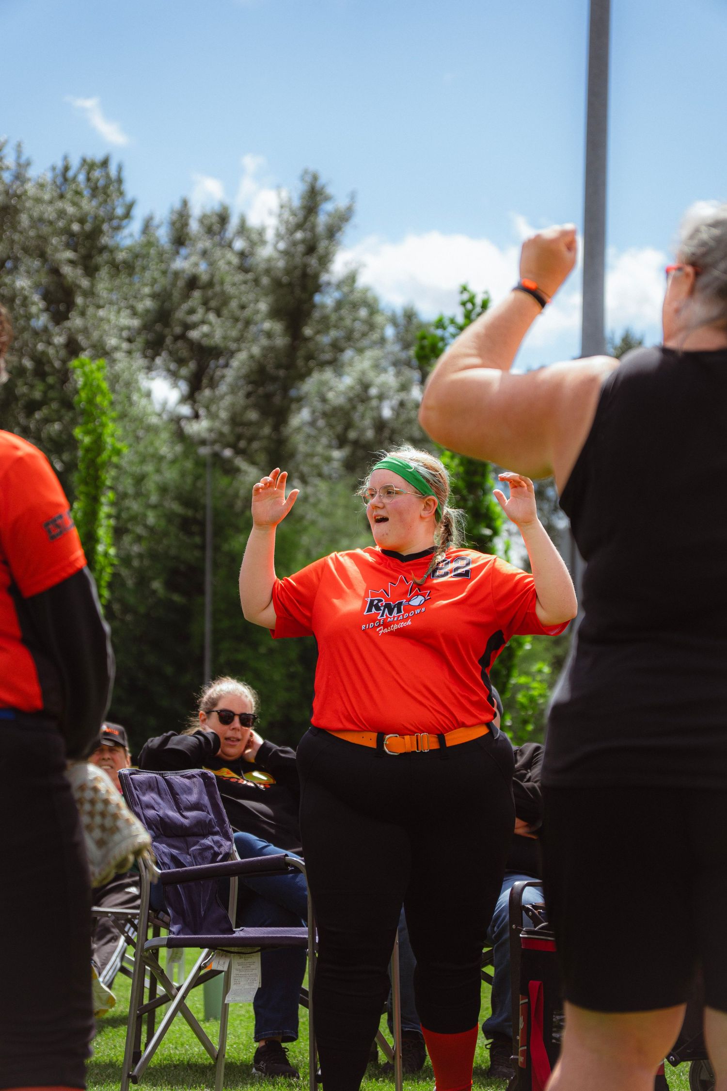 Enthusiastic female softball player in orange jersey celebrating with raised hands during sunny game.