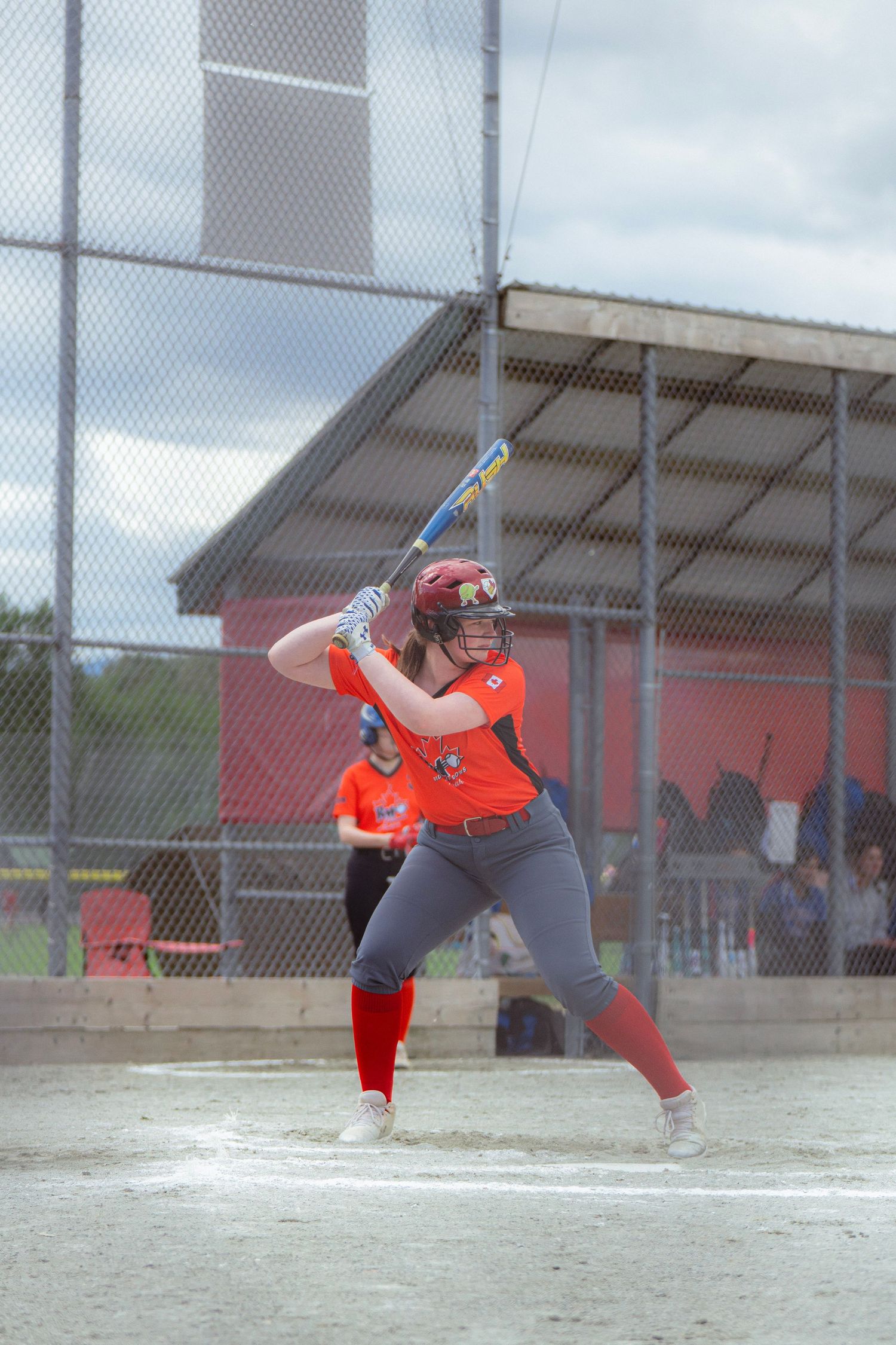 Female softball player in orange and gray uniform stands in batting stance at home plate.