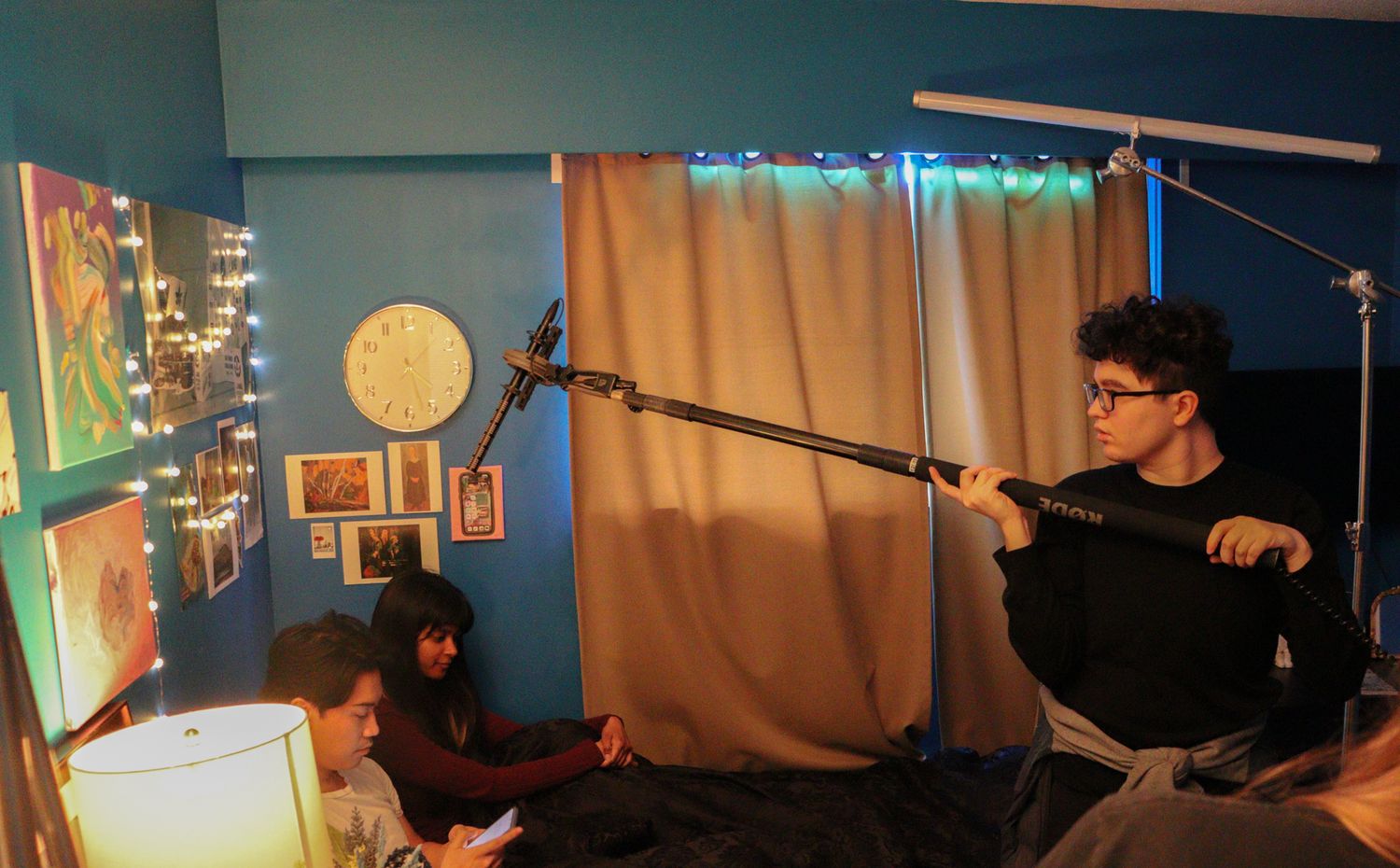 Boom operator holding a microphone over actors on a bed during an indoor film production.
