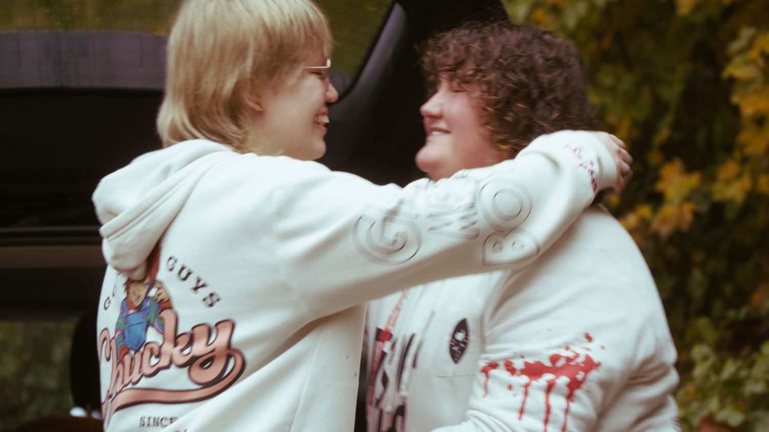 Two people in matching white graphic hoodies smile and embrace near a vehicle in autumn.