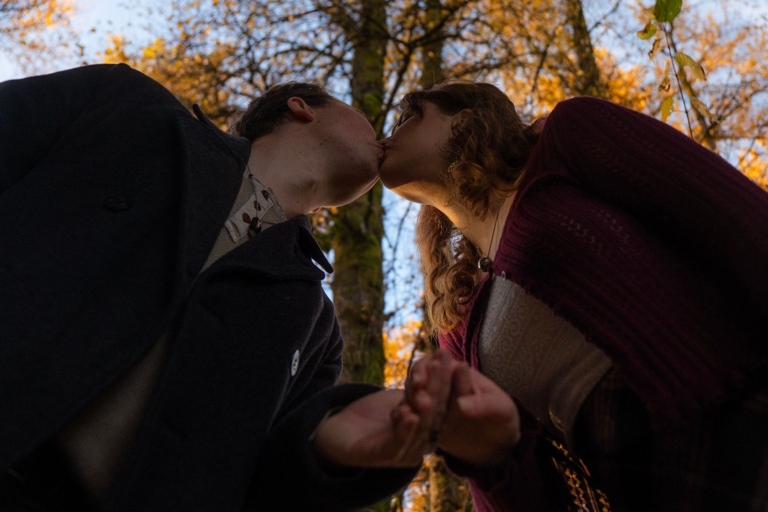 Couple sharing a kiss in autumn forest, holding hands, low angle shot with golden foliage above.
