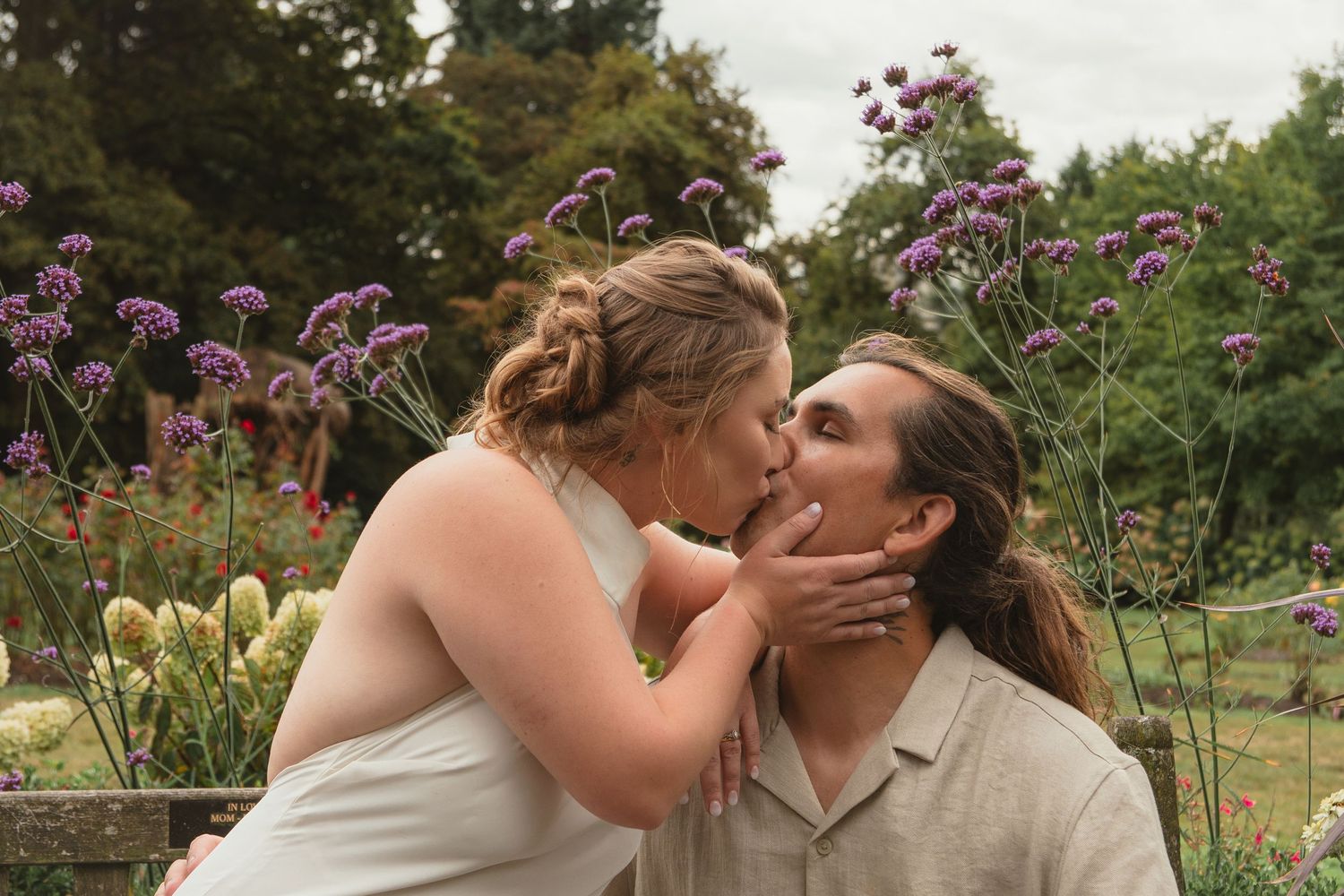 Couple kissing in a lush garden surrounded by purple verbena flowers on a cloudy day.