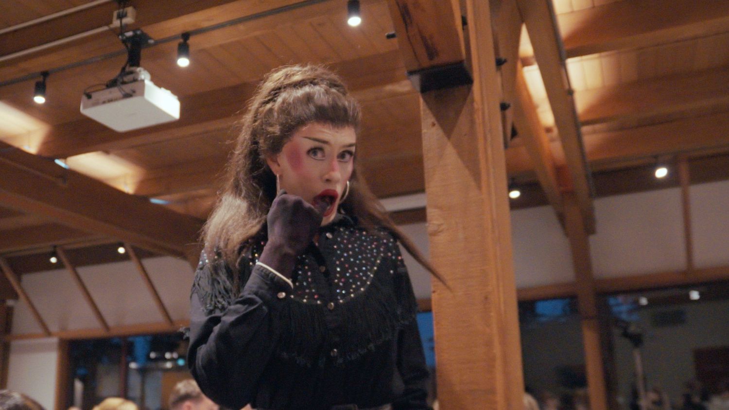 Woman in black studded jacket with gloves posing indoors under wooden ceiling beams.