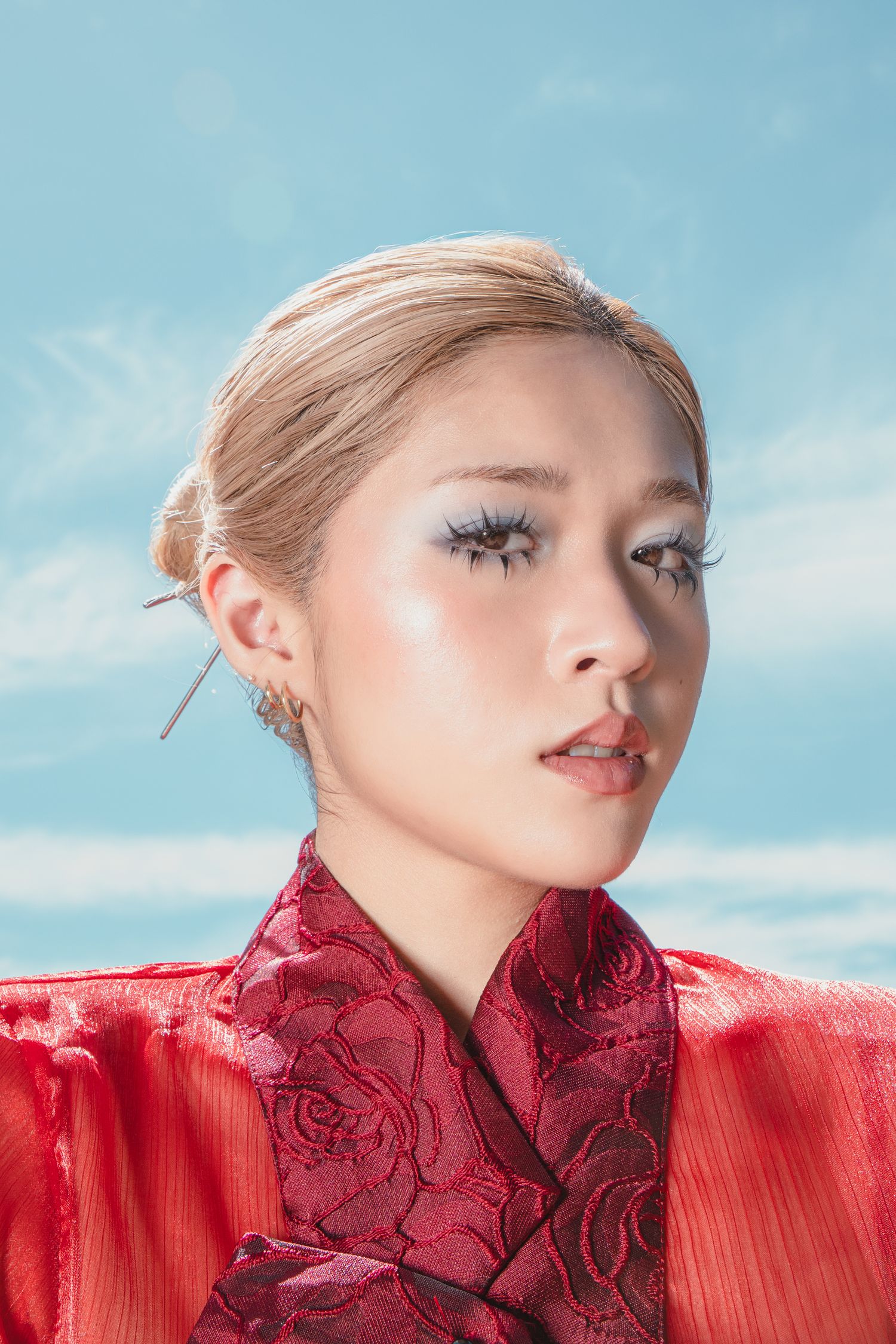 Young Asian woman in red floral cheongsam with dramatic lashes and hair pin against blue sky.