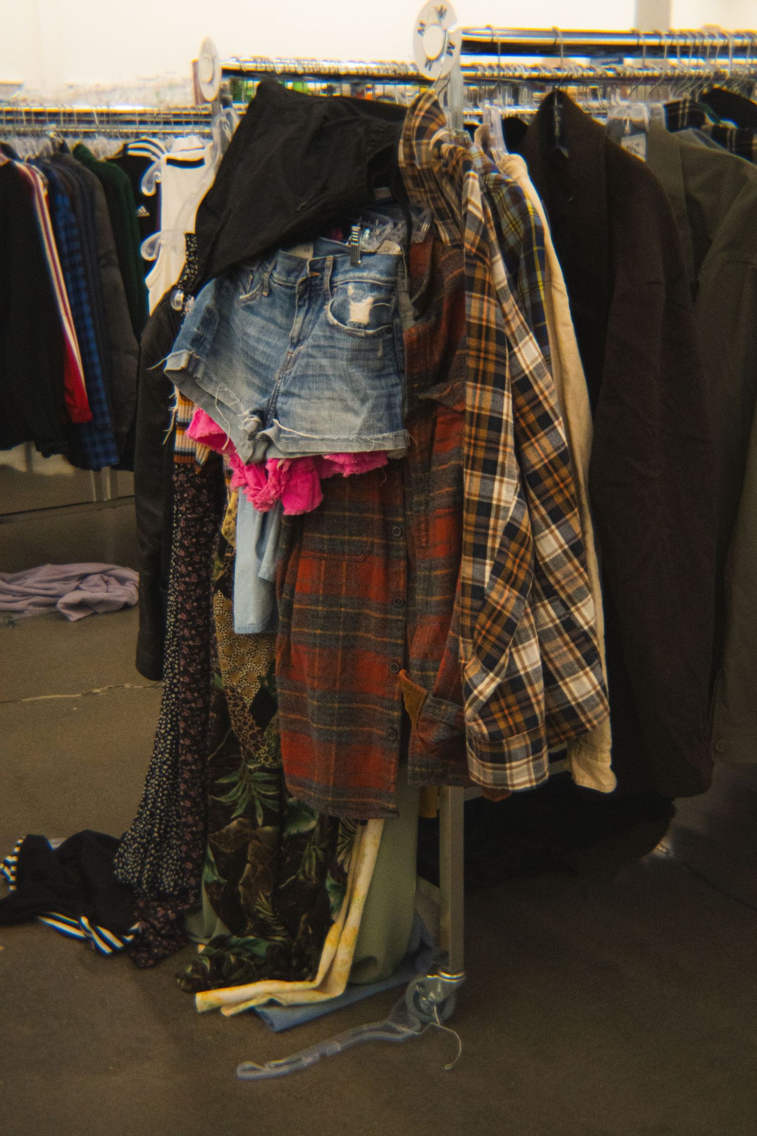 Assorted secondhand clothing including denim shorts and plaid shirts on a thrift store rack.