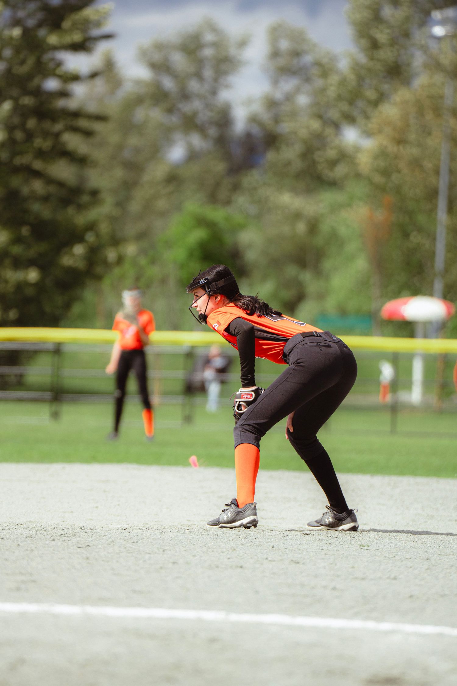 Female softball infielder in orange uniform crouches in ready position on base during outdoor game.