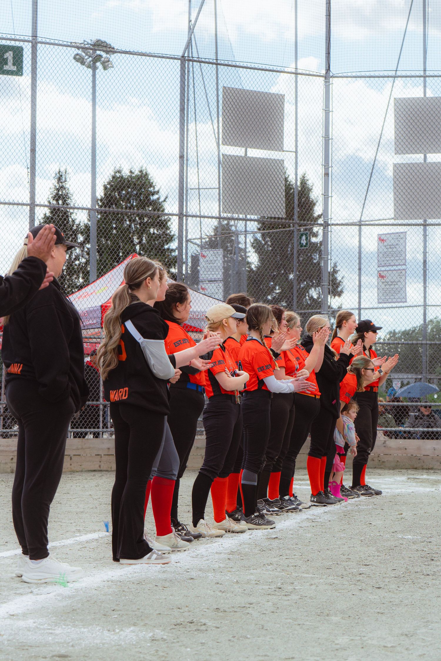 Female softball team in orange uniforms clapping together along baseline during ceremony.