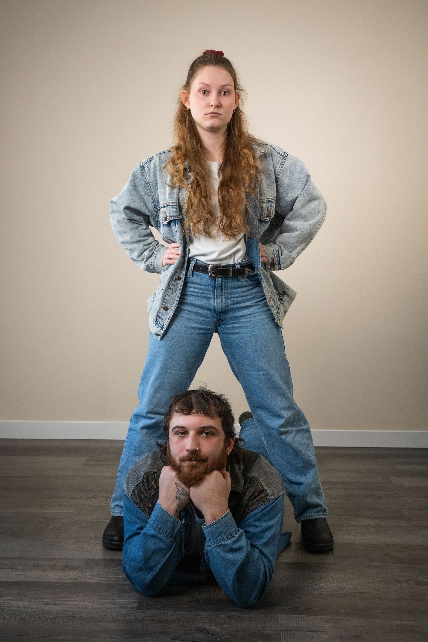 Woman in acid-wash denim jacket stands confidently over a bearded man lying on the floor in matching denim.