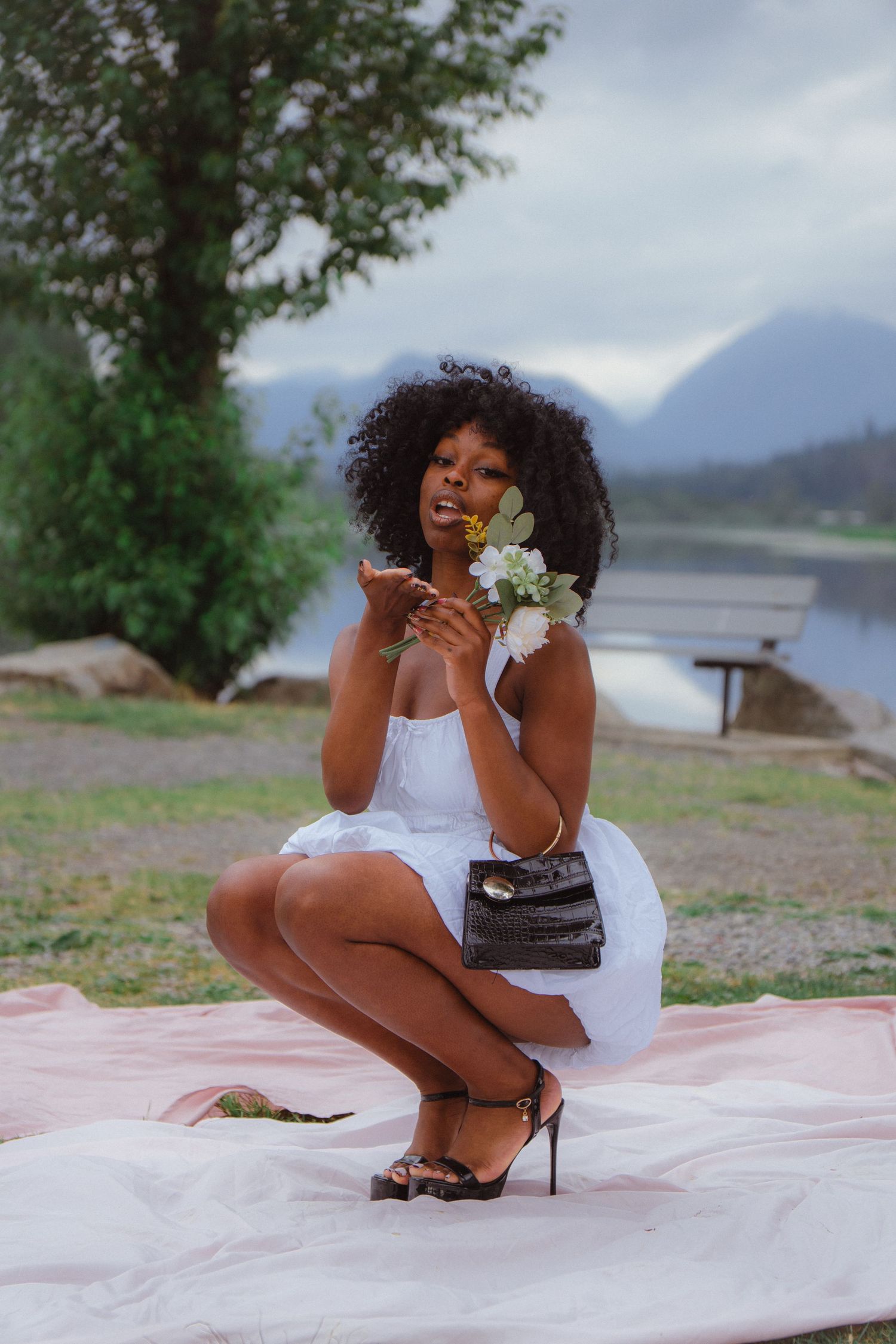 Black woman in white dress crouching with flowers and purse on pink blanket by mountain lake.