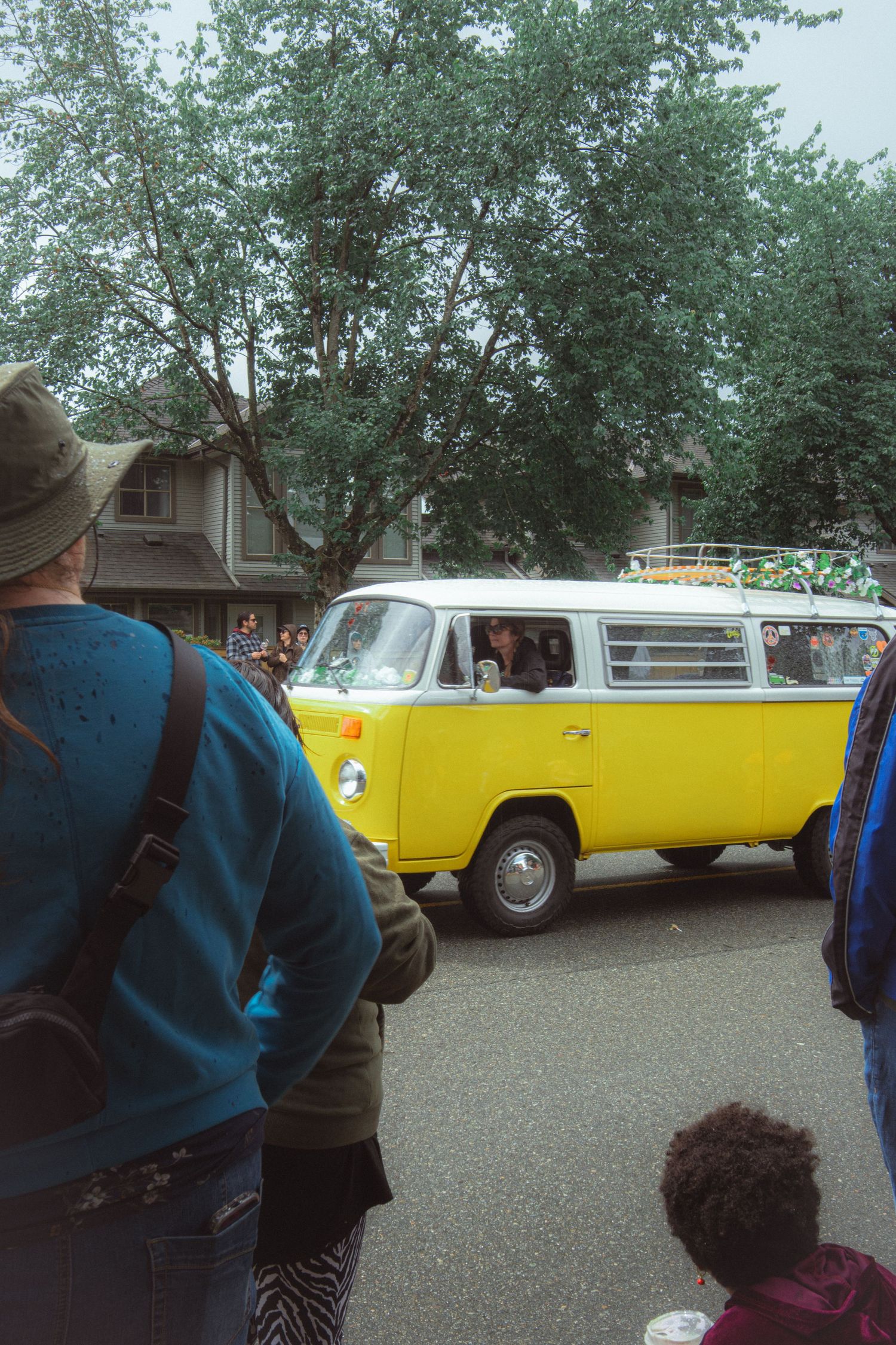 Spectators watch a vintage yellow and white VW Bus drive past during an outdoor street parade.