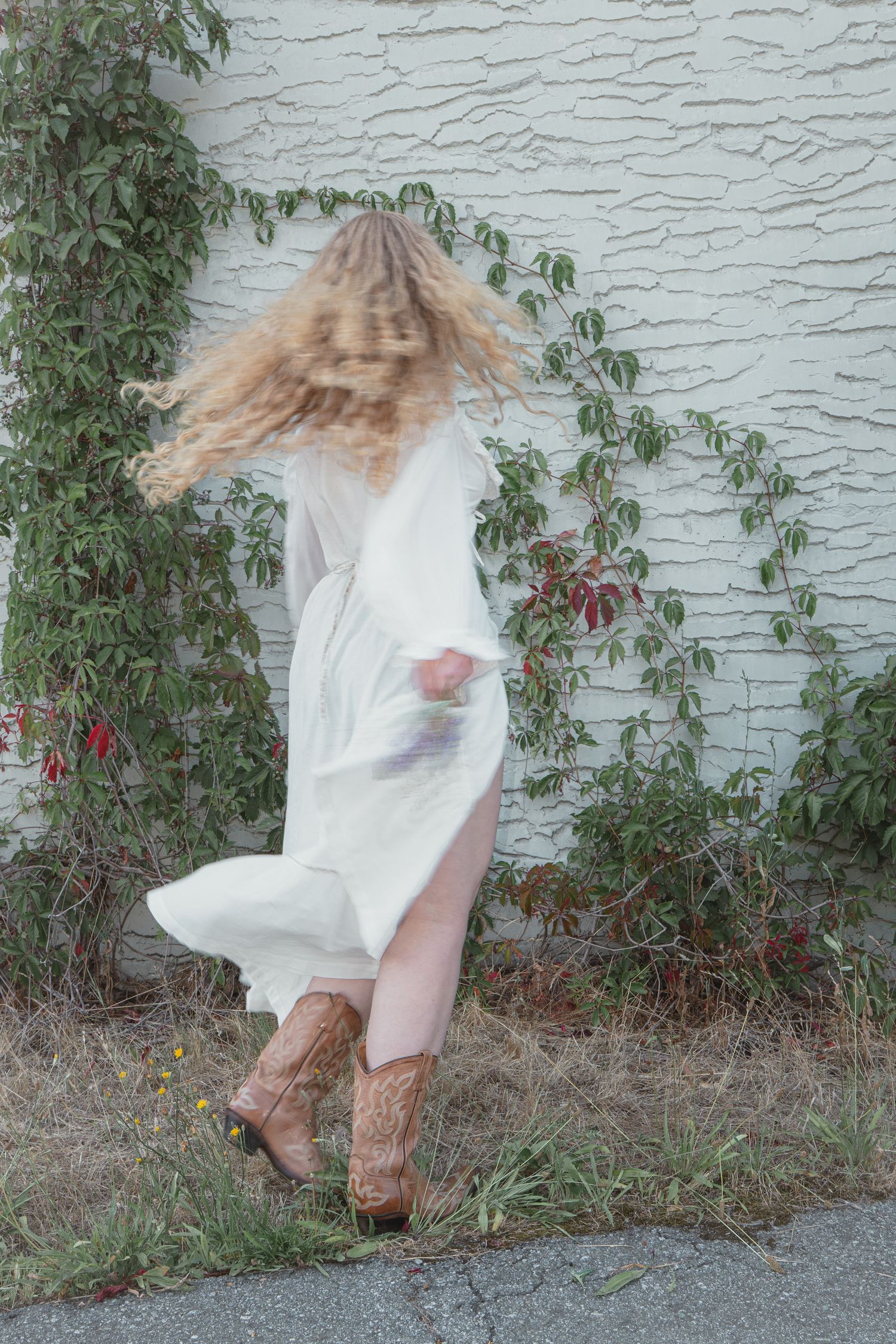 Blonde woman in flowing white dress and cowboy boots spinning outdoors near ivy-covered white brick wall.