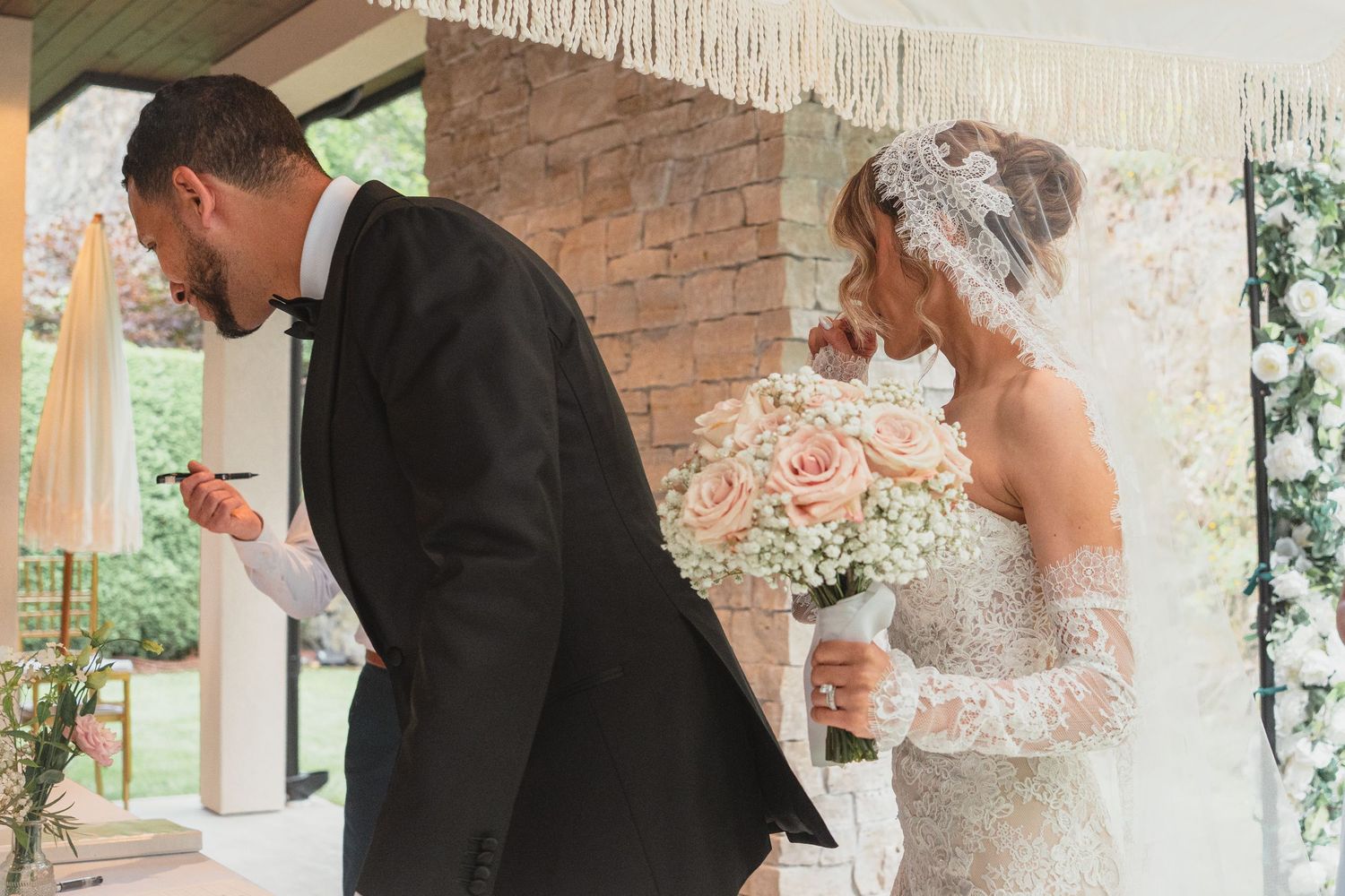 Bride holding pink rose bouquet and groom reading vows during outdoor wedding ceremony.