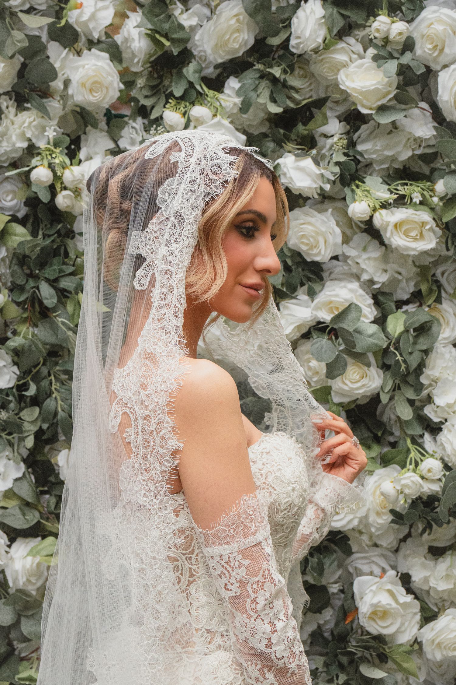 Bride in lace wedding dress and veil poses before white rose floral wall backdrop.